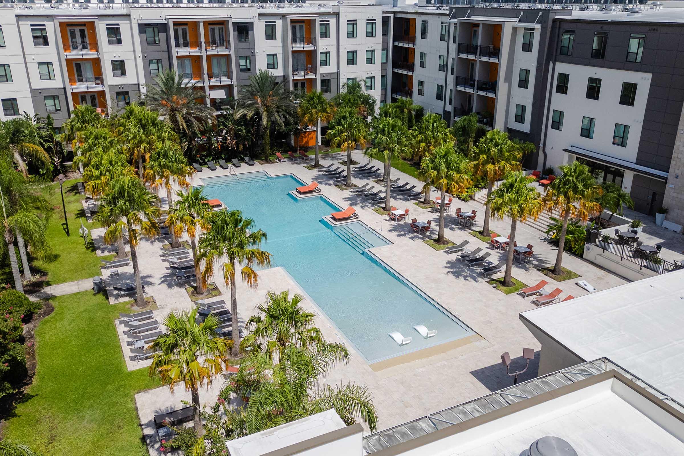 Aerial view of a modern apartment complex featuring a central swimming pool surrounded by palm trees. Lounge chairs line the pool area, and additional seating is available nearby. The buildings have a contemporary design, with multiple stories and balconies, set in a vibrant, tropical landscape.