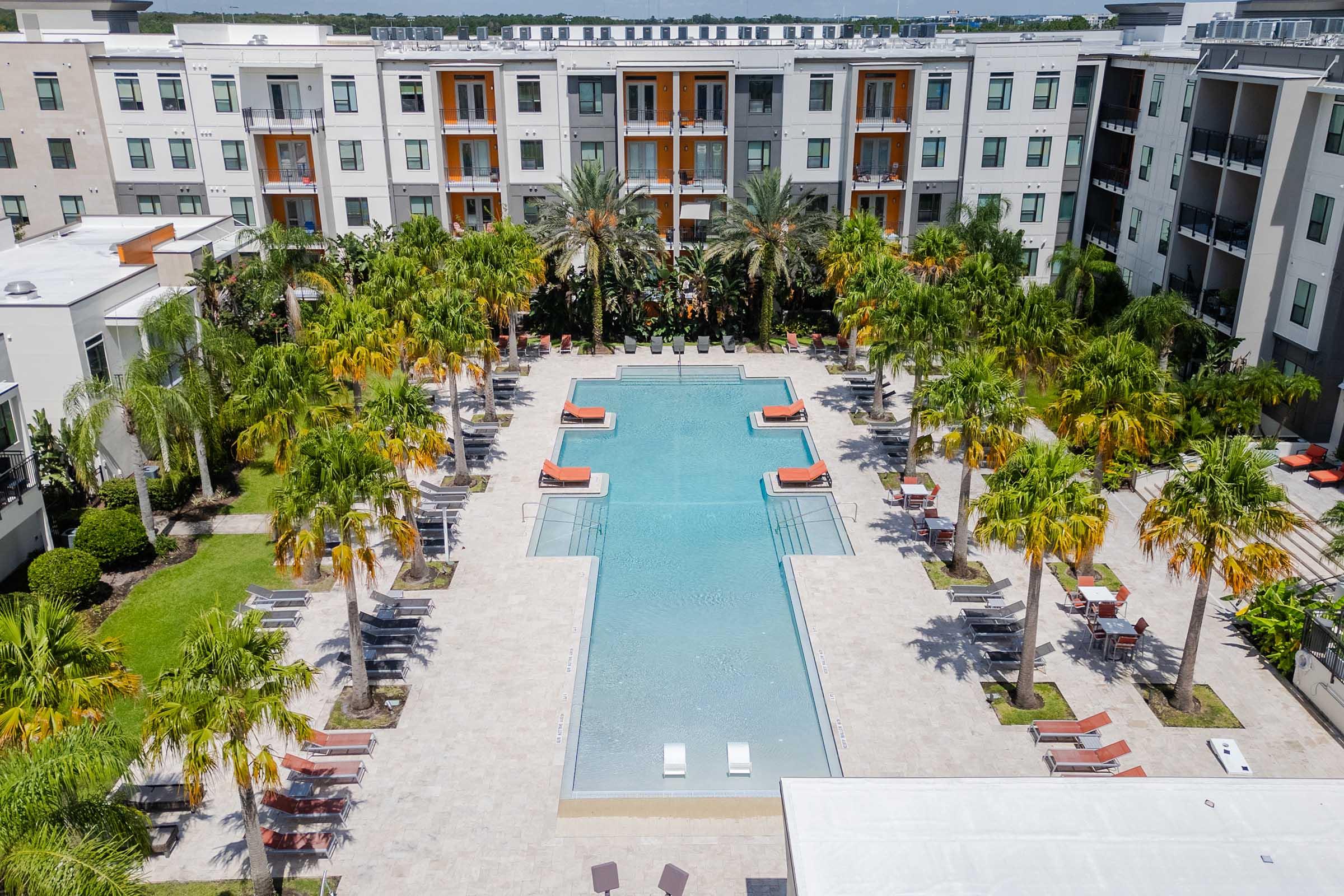 Aerial view of a modern apartment complex featuring a large swimming pool surrounded by palm trees. Lounge chairs are arranged along the poolside, and the building in the background has multiple stories with balconies. The scene is set in a sunny environment, creating a relaxed atmosphere.