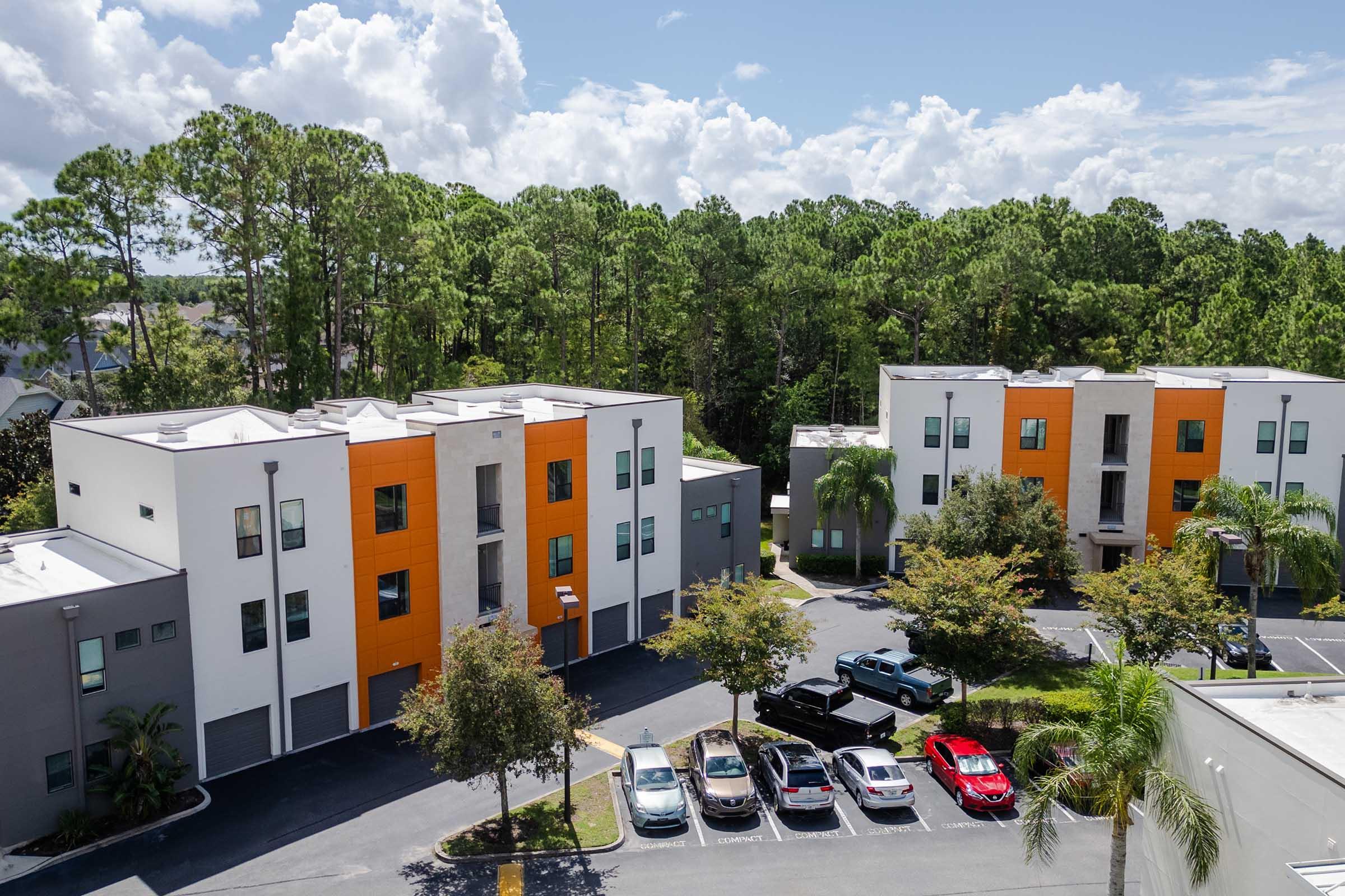 Aerial view of modern multi-family apartment buildings with orange and gray accents, surrounded by lush green trees. Several parked cars are visible in the foreground, and a clear blue sky with scattered clouds is seen above.