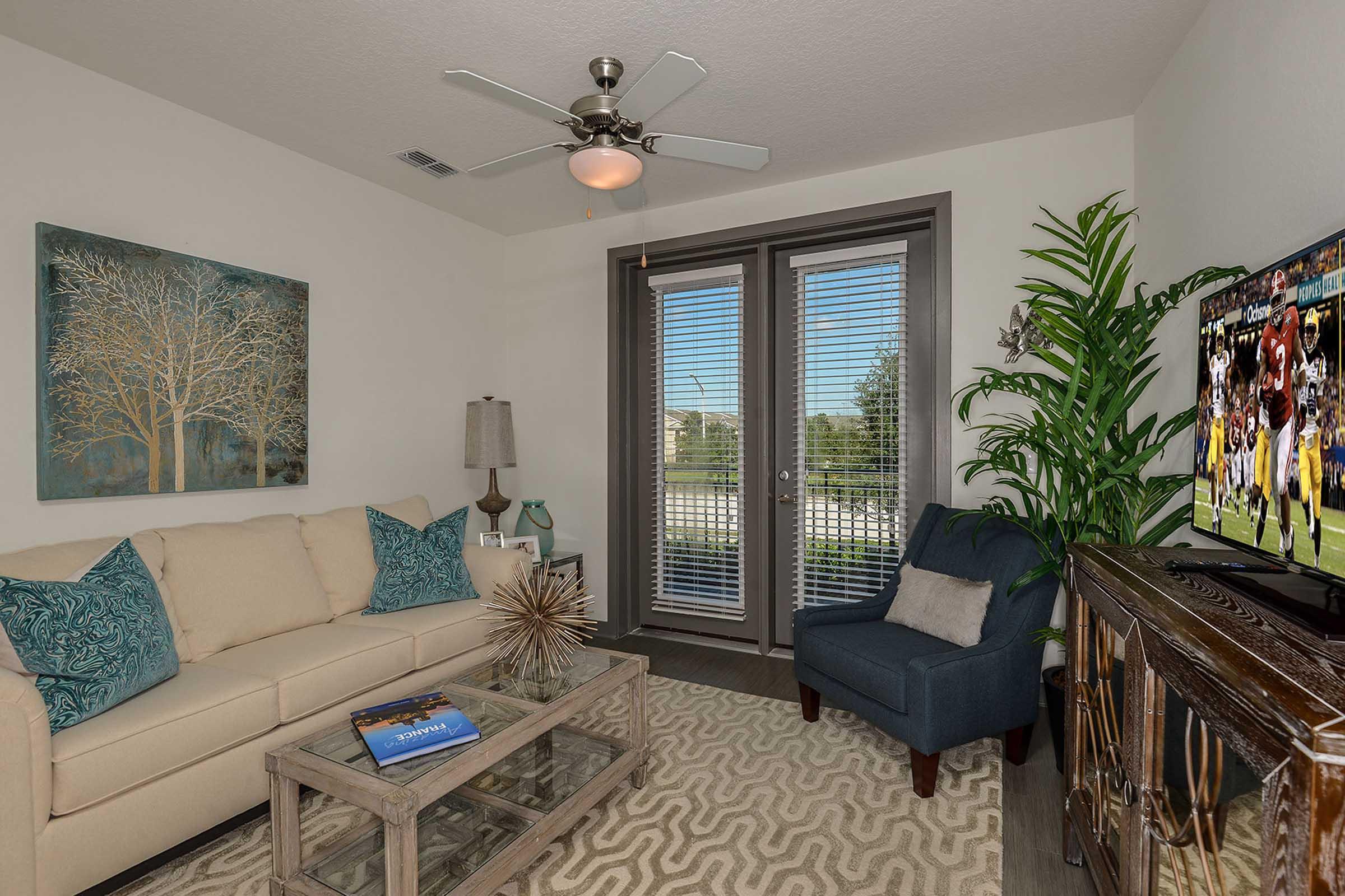 A cozy living room featuring a light-colored sofa with decorative pillows, a modern accent chair, a glass coffee table, and a TV on a wooden console. Decorative artwork hangs on the wall, and a potted plant adds greenery. Large windows with blinds allow natural light to flood the space.