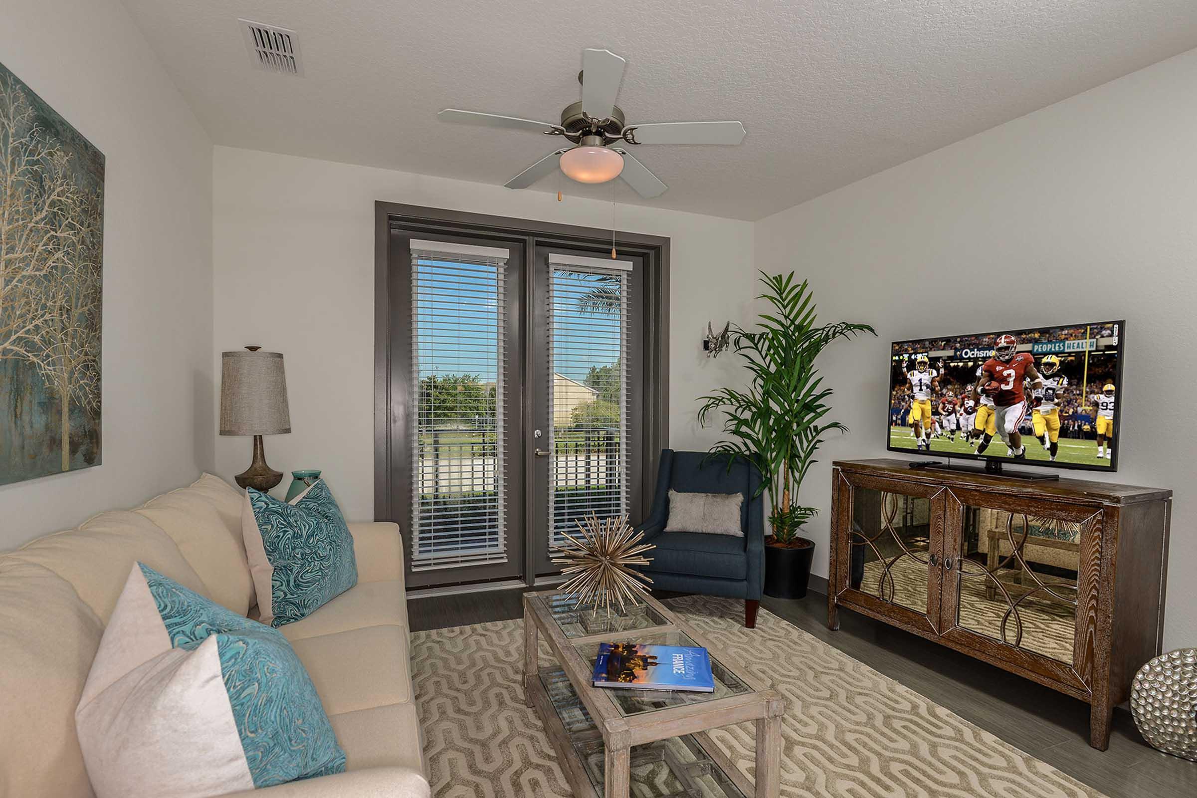 A cozy living room featuring a light beige sofa with teal throw pillows, a decorative coffee table, a stylish cabinet, and a potted plant. Natural light streams through large windows with blinds, and a flat-screen TV displays a sports game. Modern decor creates a welcoming atmosphere.