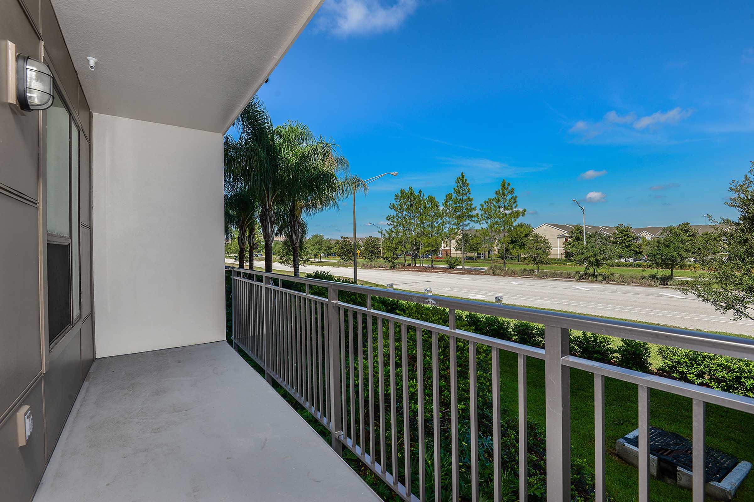 A view from a balcony with a railing, overlooking a landscaped area and a street. In the background, there are trees and buildings against a clear blue sky. The scene evokes a sense of openness and tranquility.