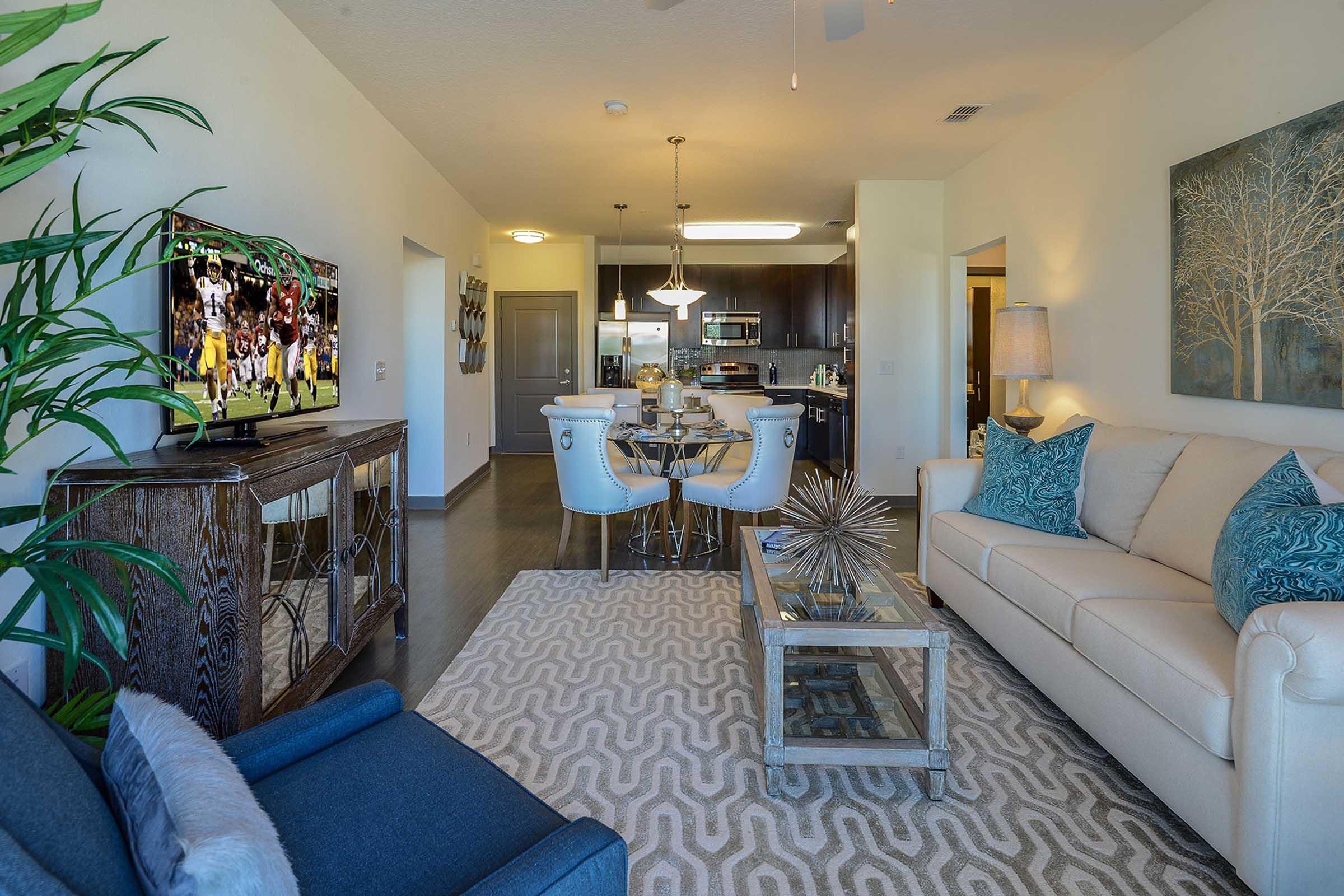 A modern living room featuring a light-colored sofa with blue accent pillows, a glass coffee table, and a patterned area rug. There is a flat-screen TV mounted on the wall, a dining area with white chairs, and a kitchen in the background. Decorative plants and artwork add a touch of warmth to the space.