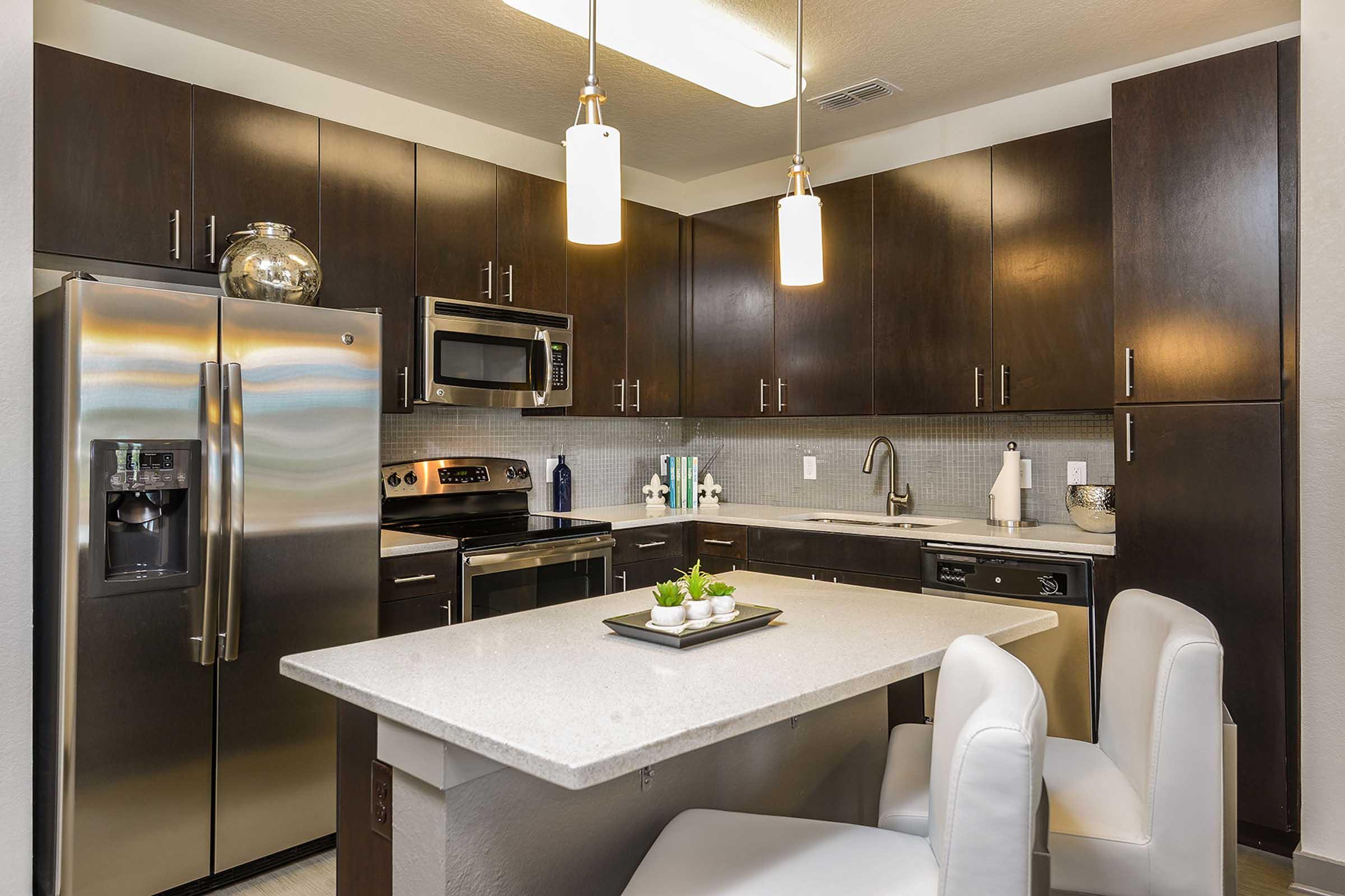 A modern kitchen featuring dark wood cabinets, stainless steel appliances including a refrigerator and oven, gray countertops, and a small dining area with white chairs and a decorative centerpiece. Bright overhead lighting enhances the contemporary design.