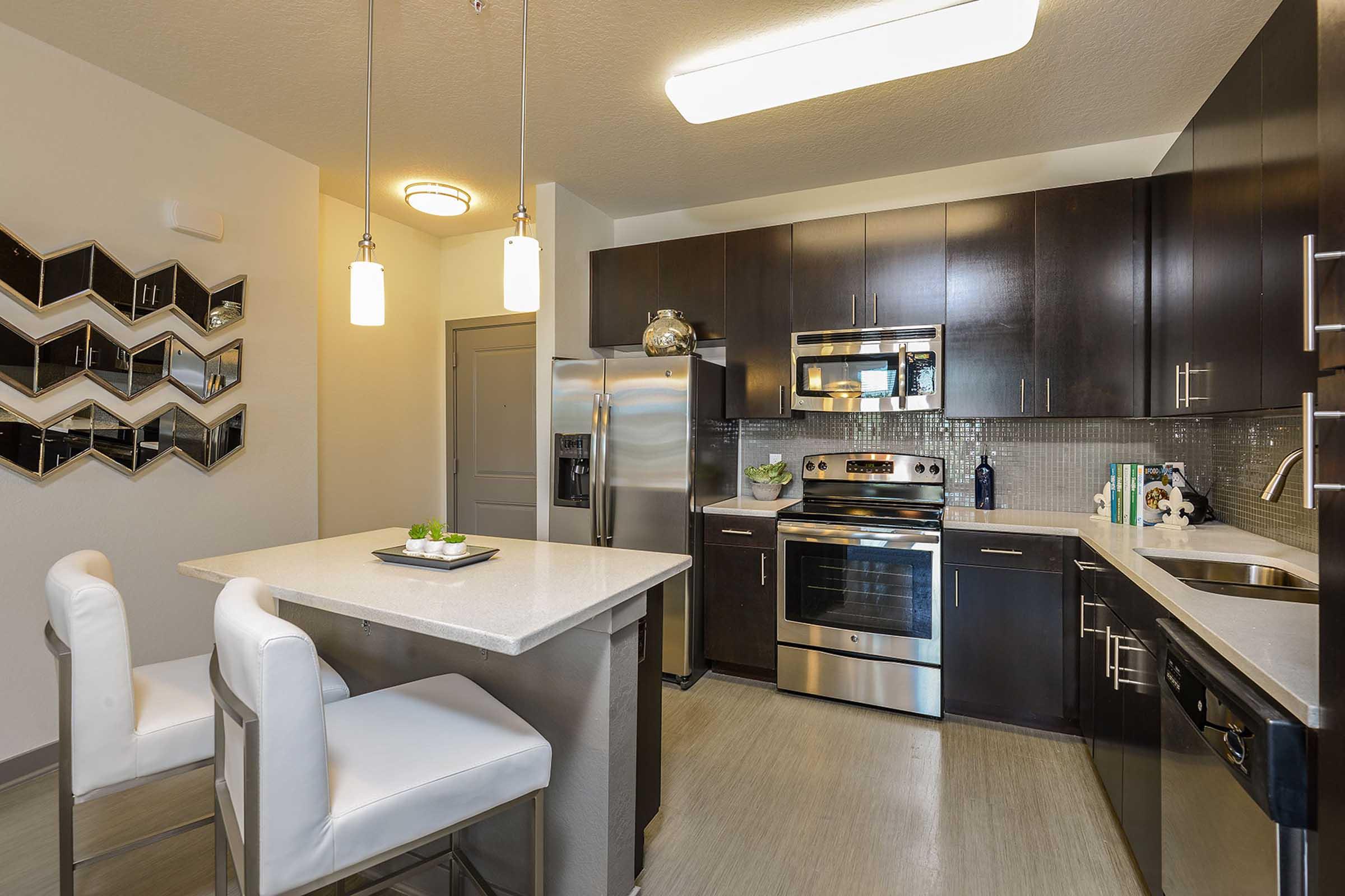 Modern kitchen featuring dark wood cabinetry, stainless steel appliances, and a stylish backsplash. A small dining area with two white chairs and a table is in the foreground. Decorative wall art can be seen on the left, adding to the contemporary aesthetic. Bright lighting enhances the warm atmosphere.