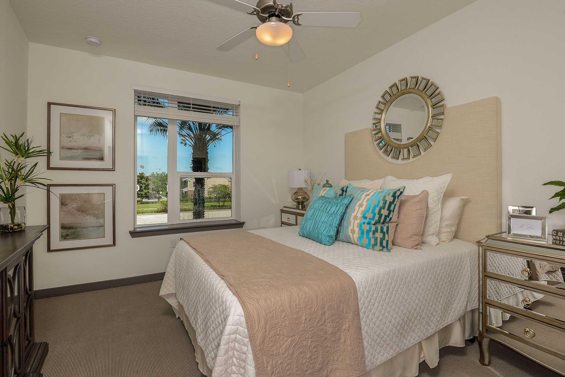 Cozy bedroom with a queen-sized bed featuring a light-colored quilt and decorative pillows in shades of blue and beige. A ceiling fan is above, and a large window offers a view of palm trees outside. The room includes a small nightstand and framed artwork on the walls, creating a serene atmosphere.