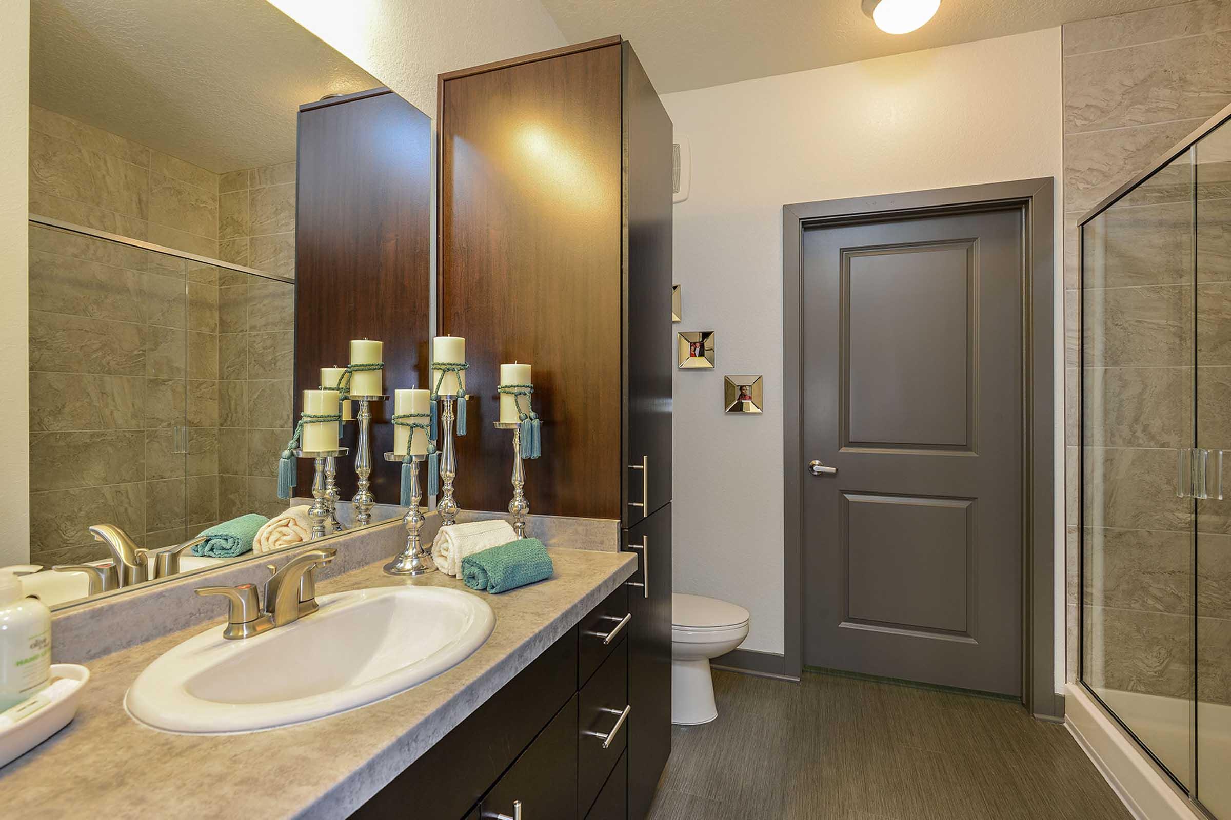 A modern bathroom featuring a vanity with a sink, mirror, and decorative candles. The space includes a shower area with glass doors, a dark gray door, and a toilet. The color scheme is neutral, with stone tile and sleek cabinetry, creating a minimalist and contemporary look.