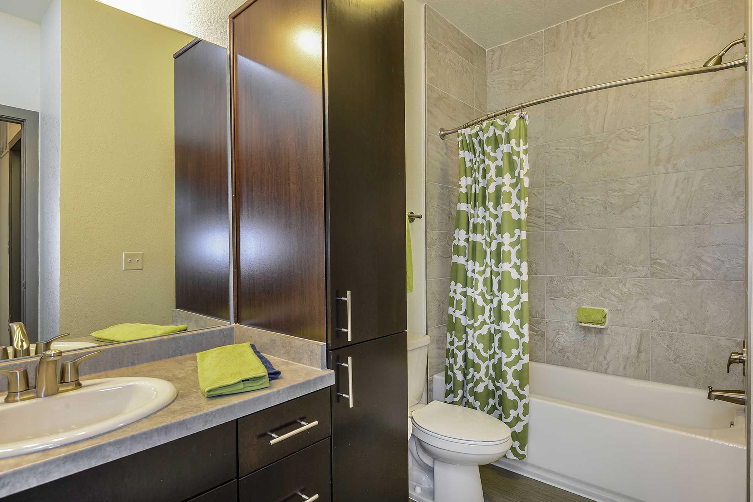 A modern bathroom featuring a white bathtub with a green and white patterned shower curtain, a dark wood vanity with a sink, and a large mirror. There are decorative towels on the counter and a lime green towel hanging on the wall. Neutral-colored tiles cover the walls and floor.