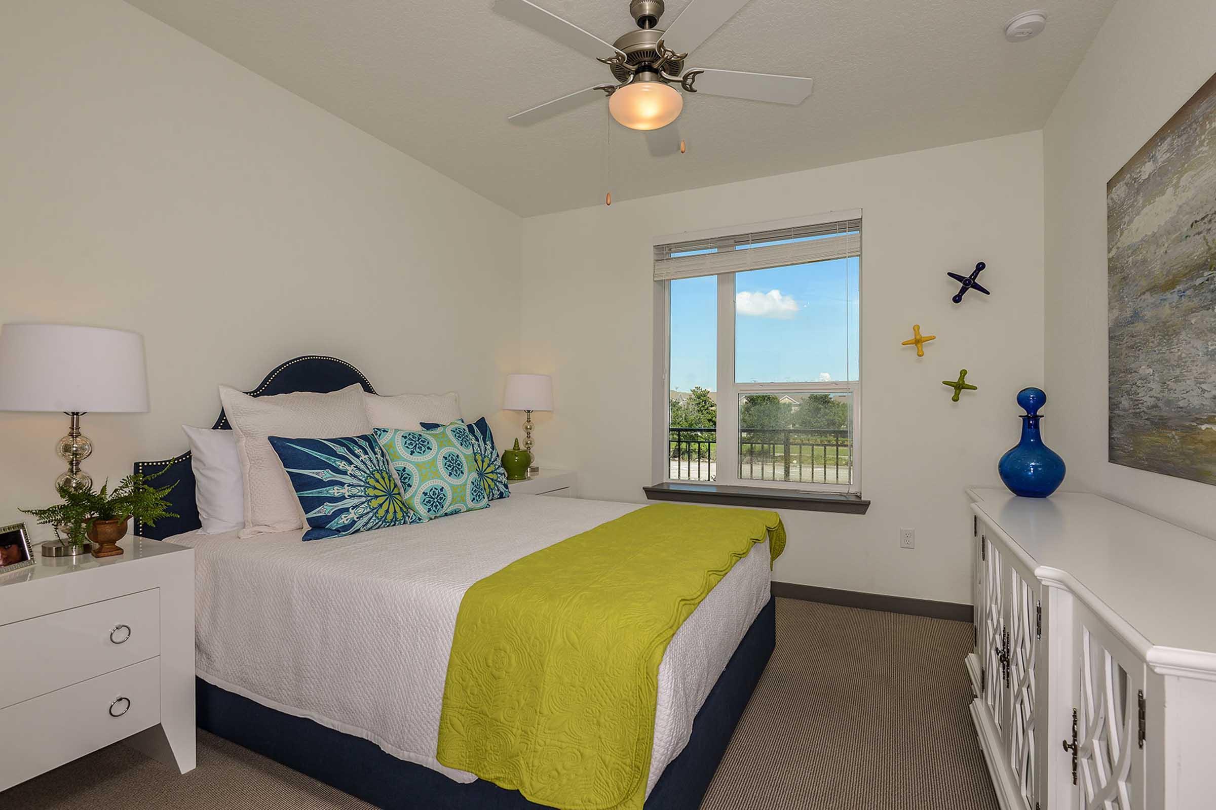 A cozy bedroom featuring a queen-sized bed with colorful pillows and a lime green throw blanket. A white nightstand with lamps on either side and a decorative blue vase. Natural light enters through a window, with a view of greenery outside. Artwork and colorful wall decorations enhance the inviting atmosphere.