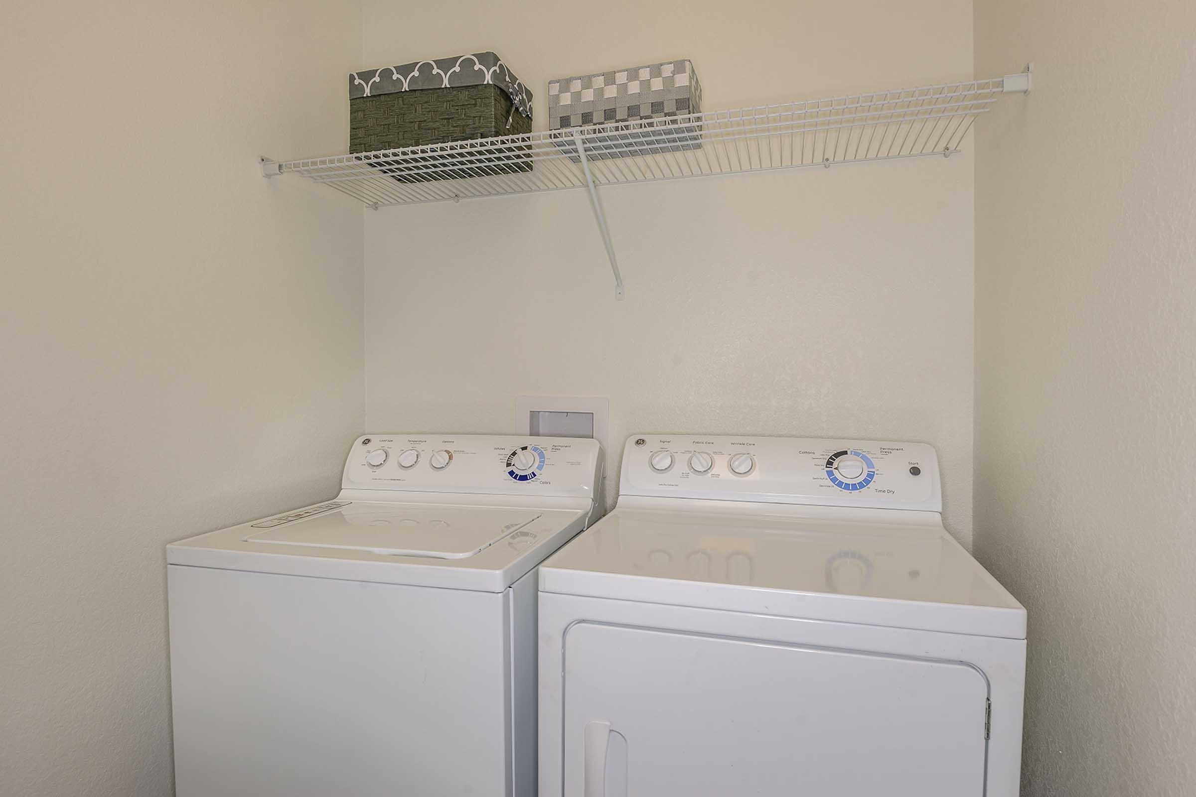 A clean laundry room featuring a white washing machine and dryer side by side. Above them, there is a white wire shelf with two decorative storage boxes. The walls are a light color, creating a bright and organized space.