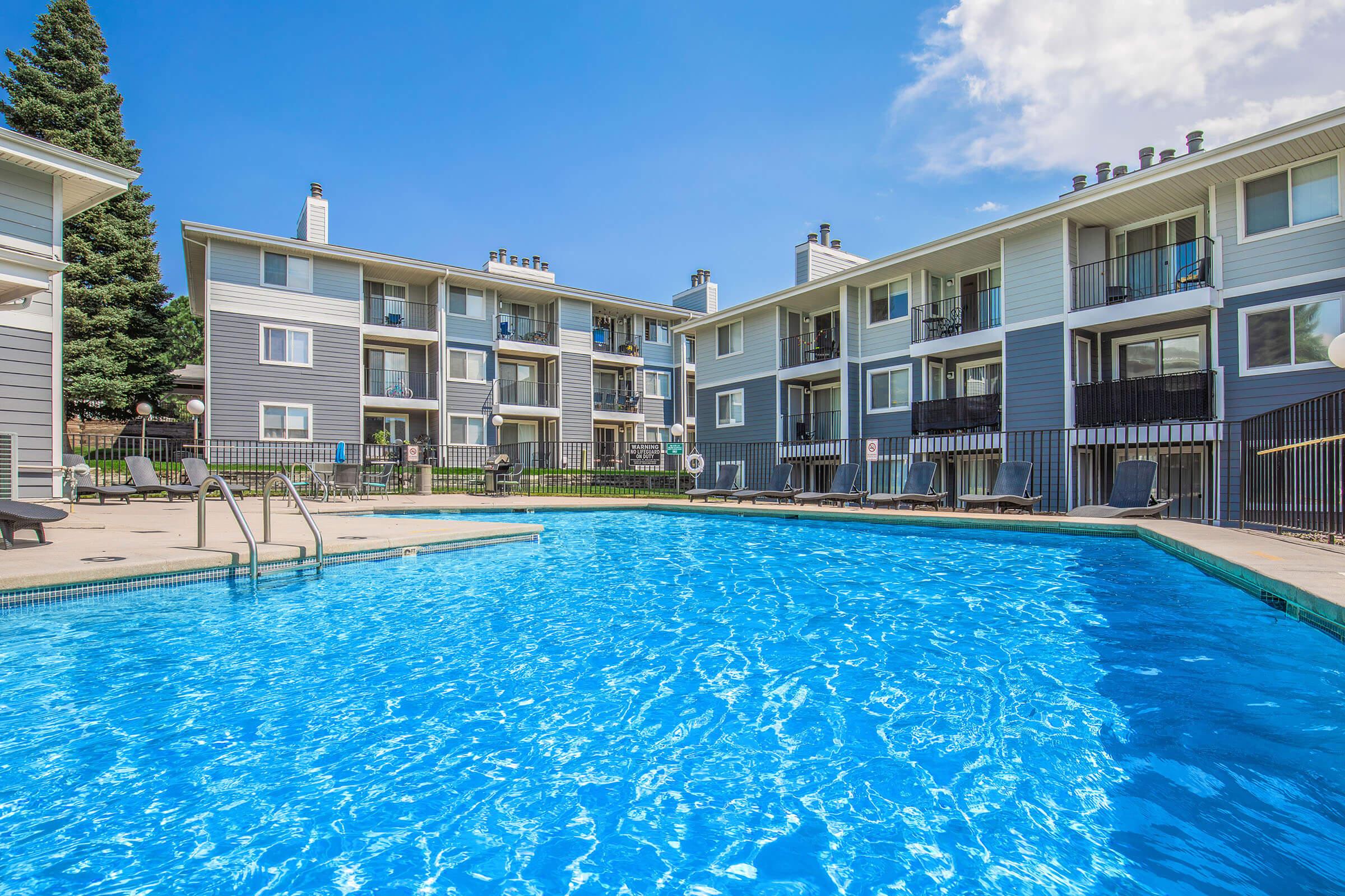 Bright blue swimming pool surrounded by a sunlit patio area with lounge chairs. In the background, there are modern, multi-story apartment buildings with balconies. Lush green trees and a clear blue sky enhance the inviting atmosphere. The scene depicts a relaxing and vibrant residential community.