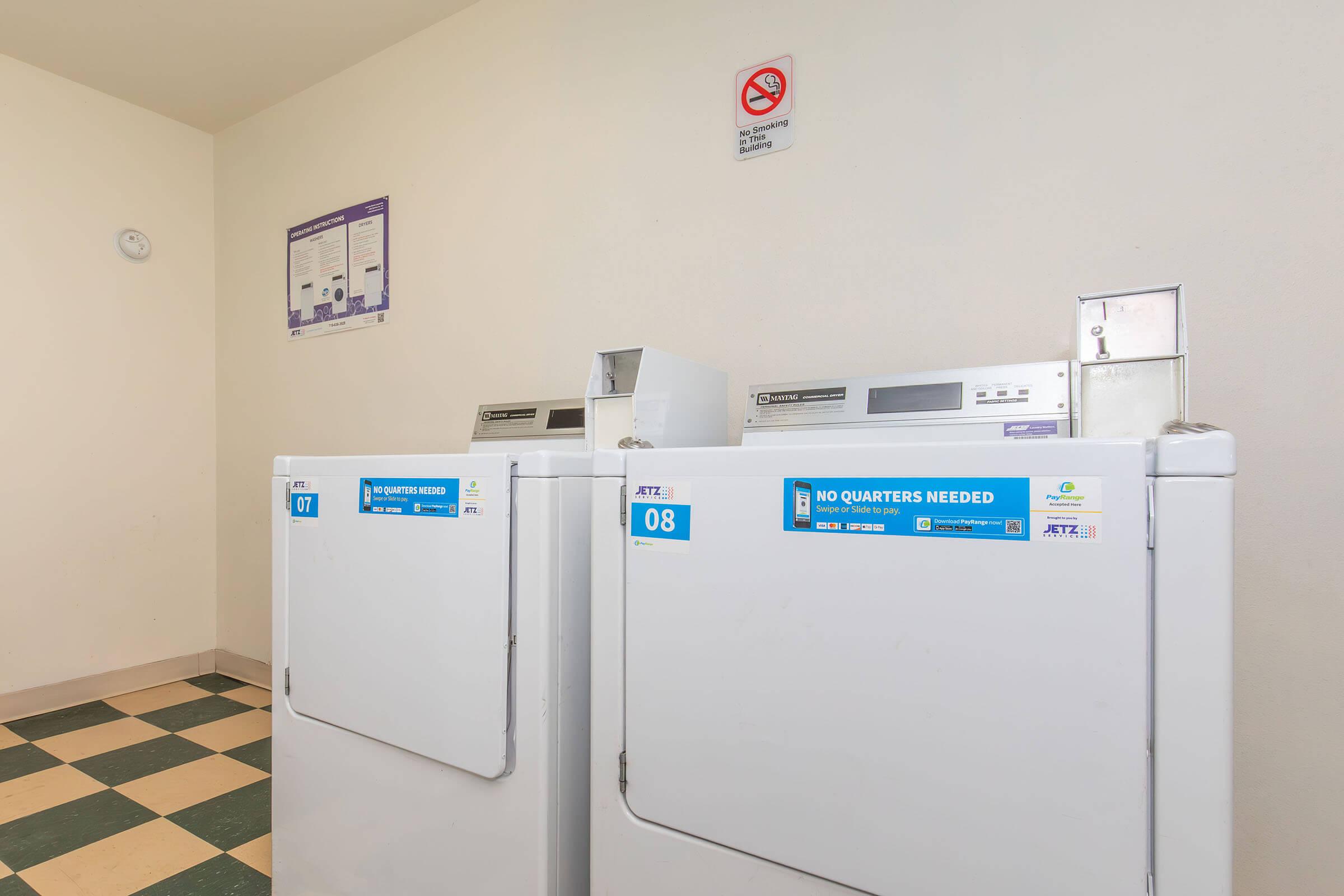 Two stacked washing machines in a laundry area, labeled with numbers 07 and 08. The machines display a sign indicating that no quarters are needed for operation. The floor features a checkered pattern in green and beige tiles, and there is a no smoking sign on the wall.
