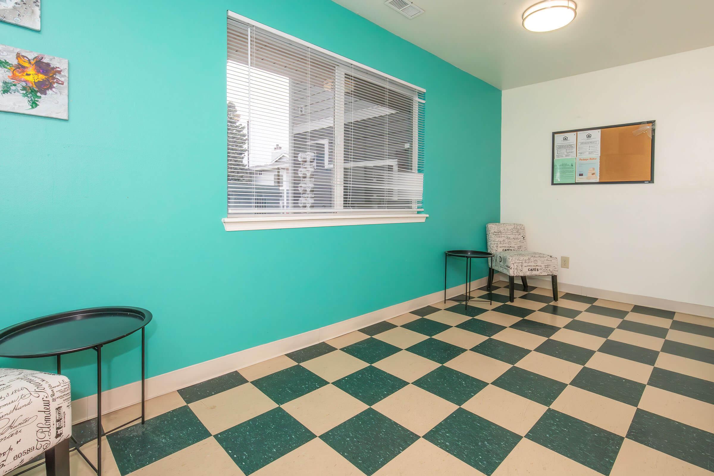 An empty room with turquoise walls and a checkered black and white floor. Two small chairs are positioned in opposite corners, alongside black metal side tables. A window with blinds allows natural light to enter, and a bulletin board is mounted on one wall.