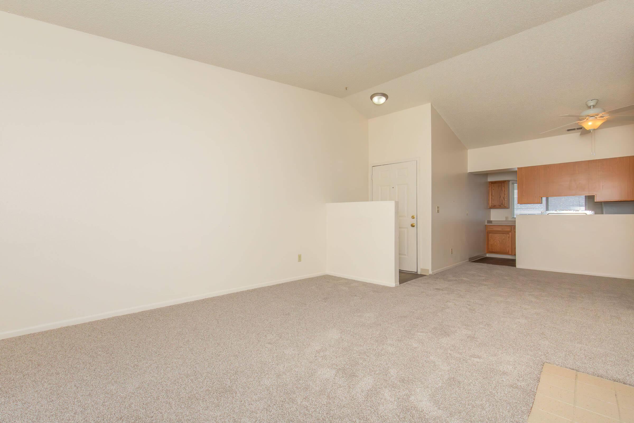 A spacious living area with beige carpeting and white walls. A doorway leads to a kitchen in the background, which features wooden cabinets. Natural light fills the room, creating a bright and airy atmosphere. The ceiling has a light fixture and a ceiling fan.