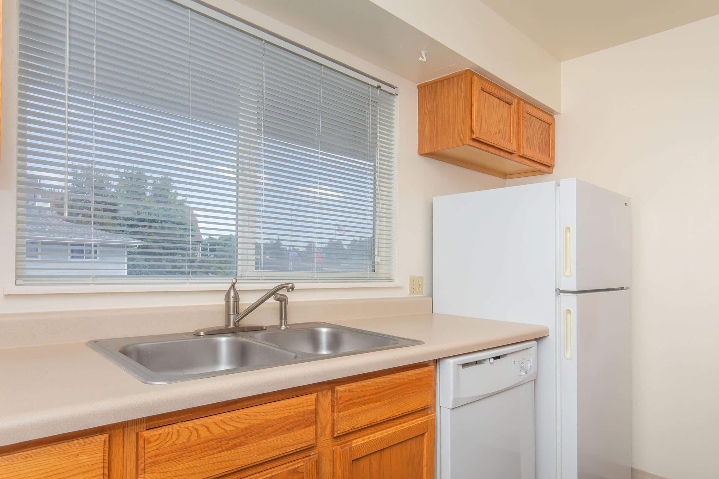 A modern kitchen featuring a double sink, light-colored countertops, wooden cabinets, a white refrigerator, and a window with blinds allowing natural light to enter. The scene is tidy and well-organized, emphasizing a functional and inviting space.