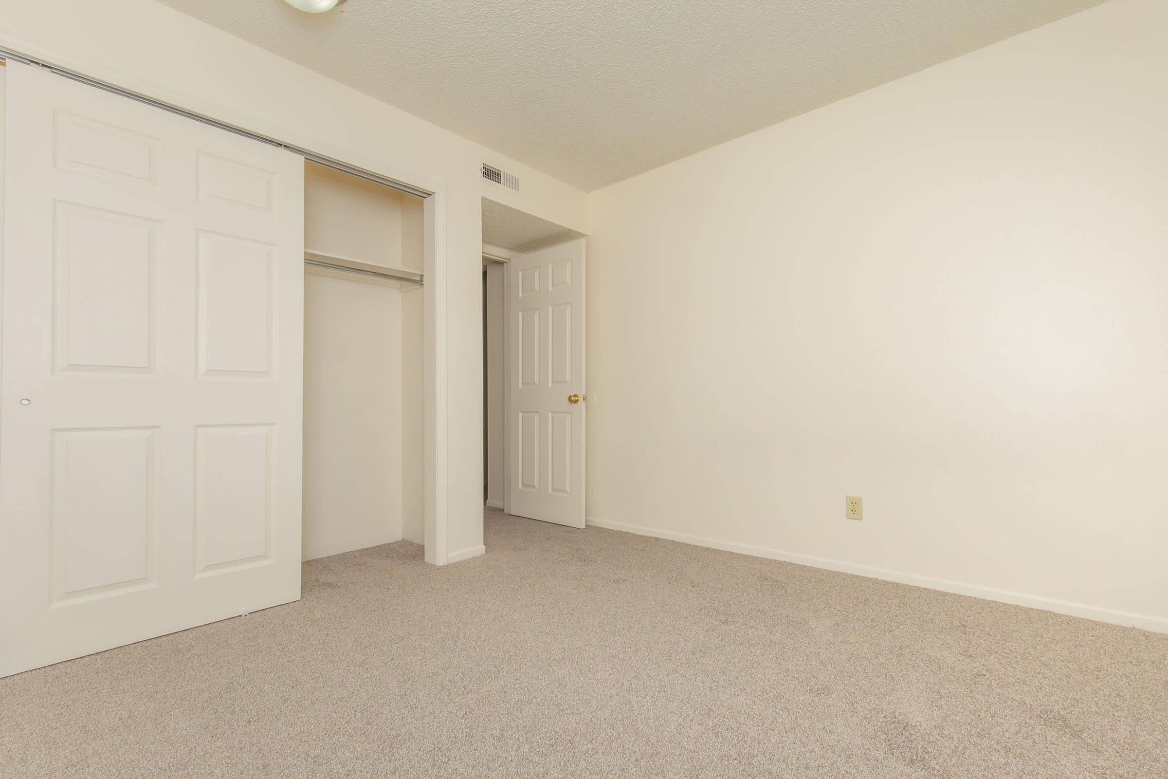 A well-lit empty room featuring light beige walls and carpet. There are two sets of closed doors: one leads to a closet with sliding doors and the other to a standard door. The space is uncluttered and has a simple, clean aesthetic. Ideal for potential customization or decoration.