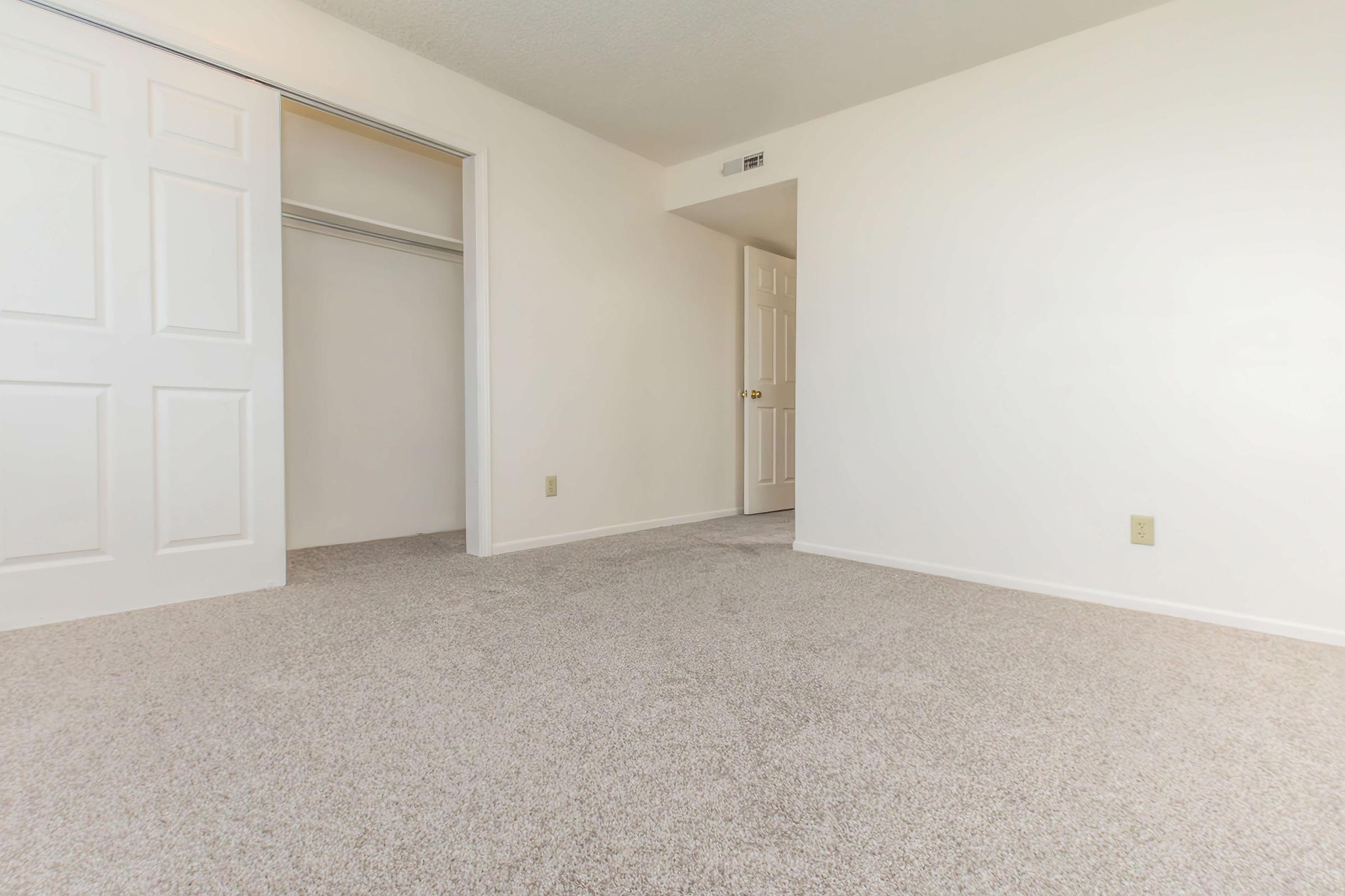 A spacious, empty room with light-colored walls and beige carpet flooring. On the left, there is an open closet with sliding doors, and on the right, a closed door leading to another room. Natural light fills the space, creating a bright and airy atmosphere.