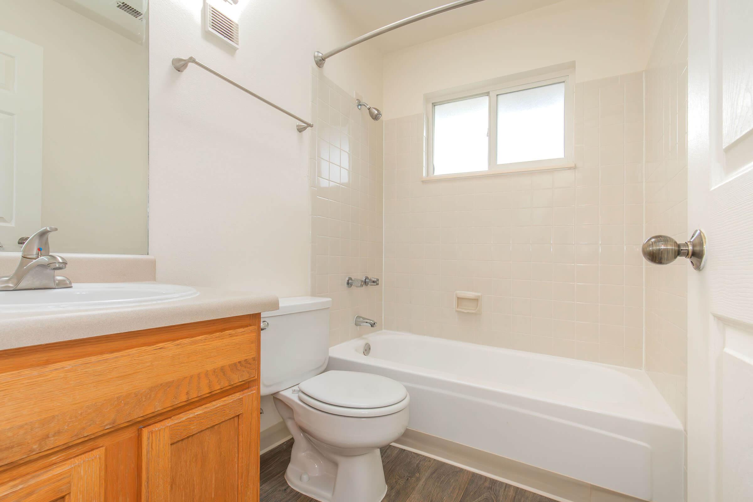 A clean and well-lit bathroom featuring a bathtub with shower, toilet, and sink. The walls are mainly tiled in light colors, and there is a window allowing natural light. The cabinetry is wooden, providing storage space.