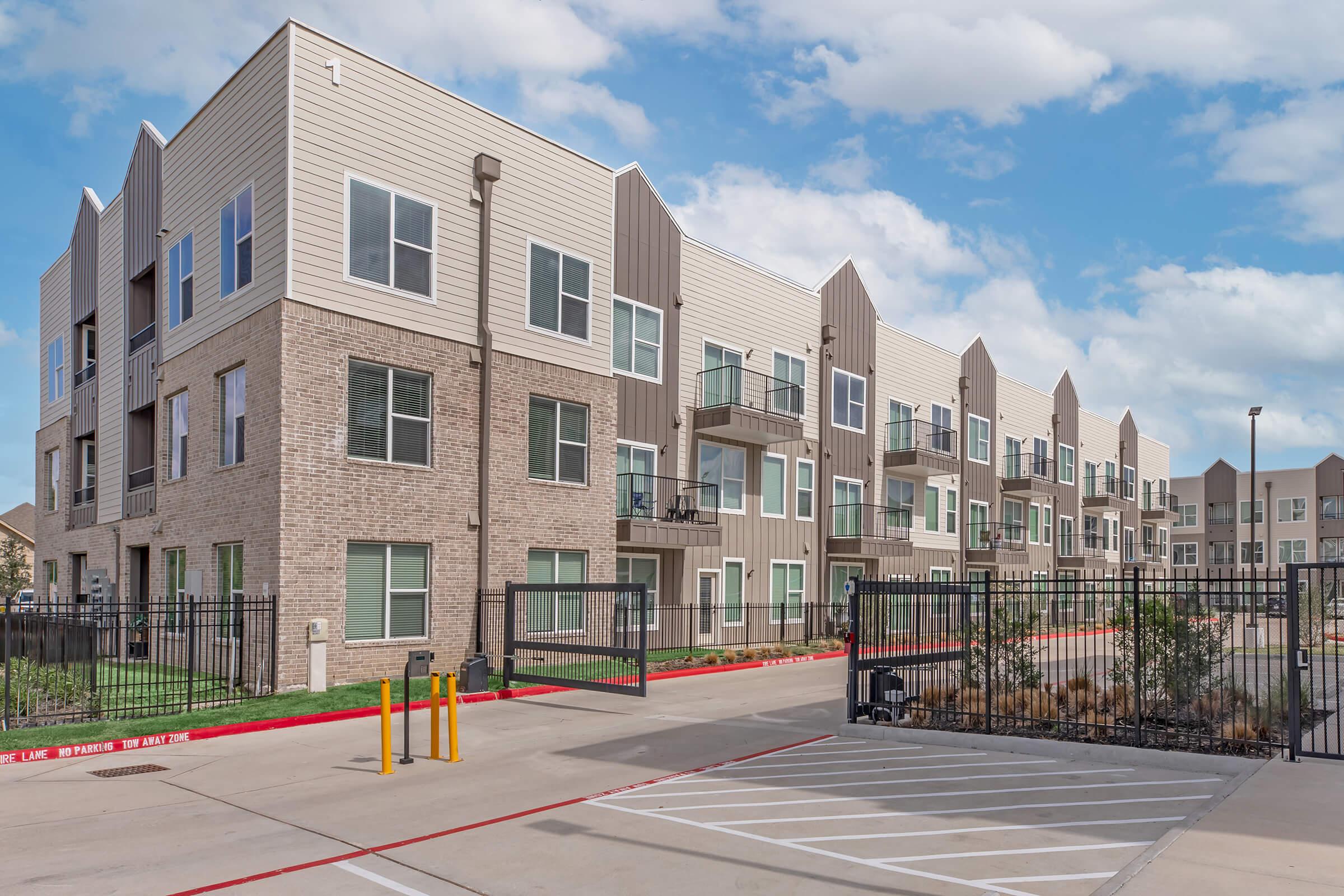 Modern apartment complex with multiple stories, featuring a combination of brick and siding exteriors. Large windows and balconies are visible. A gated entrance with yellow barrier arms is in the foreground, along with a concrete parking area and landscaped grounds under a partly cloudy sky.