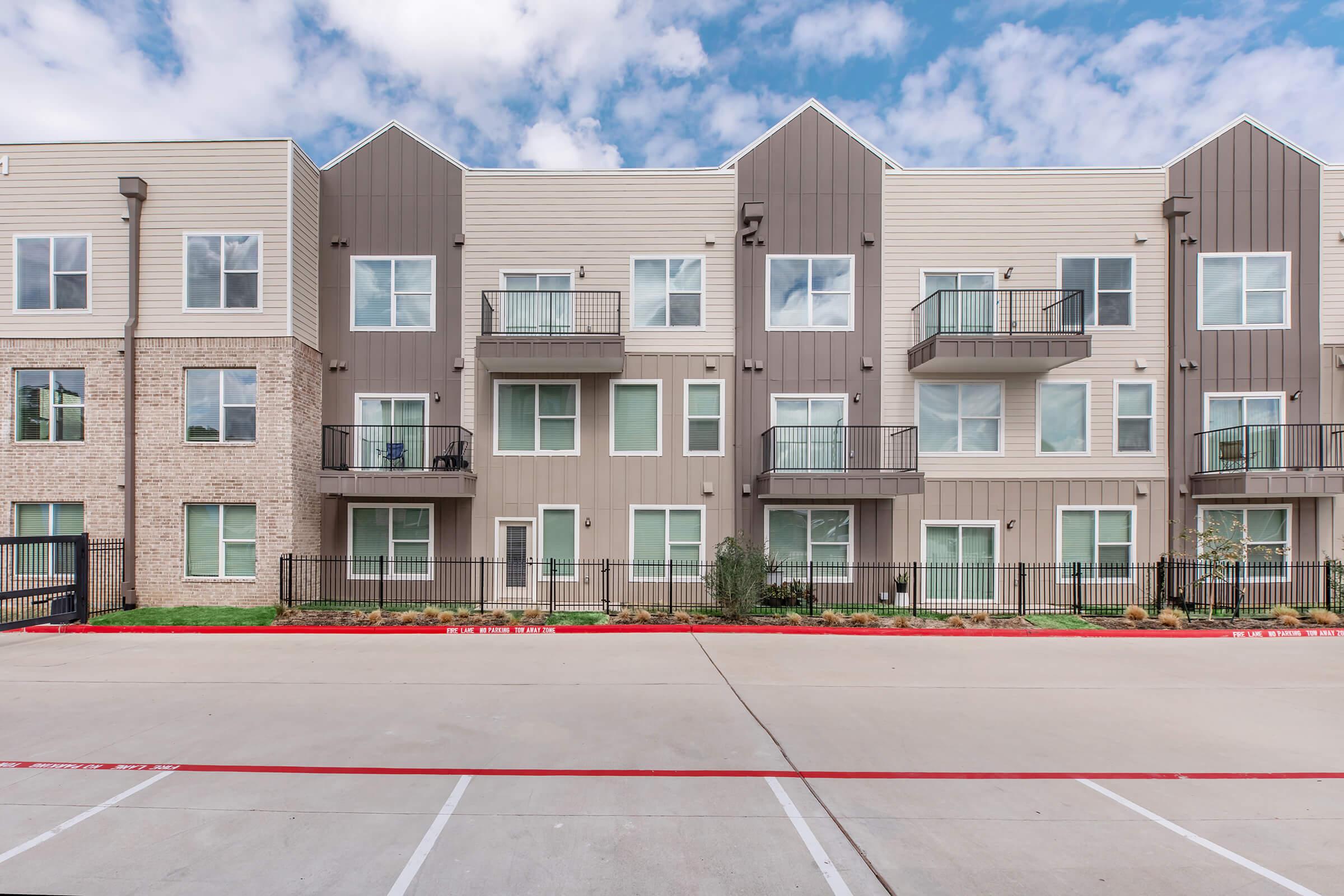 Modern multi-story apartment building with a mix of beige and brown siding. The facade features several balconies and large windows. In the foreground, there is a paved parking area with marked parking spaces. The sky above is partly cloudy, contributing to a bright atmosphere.