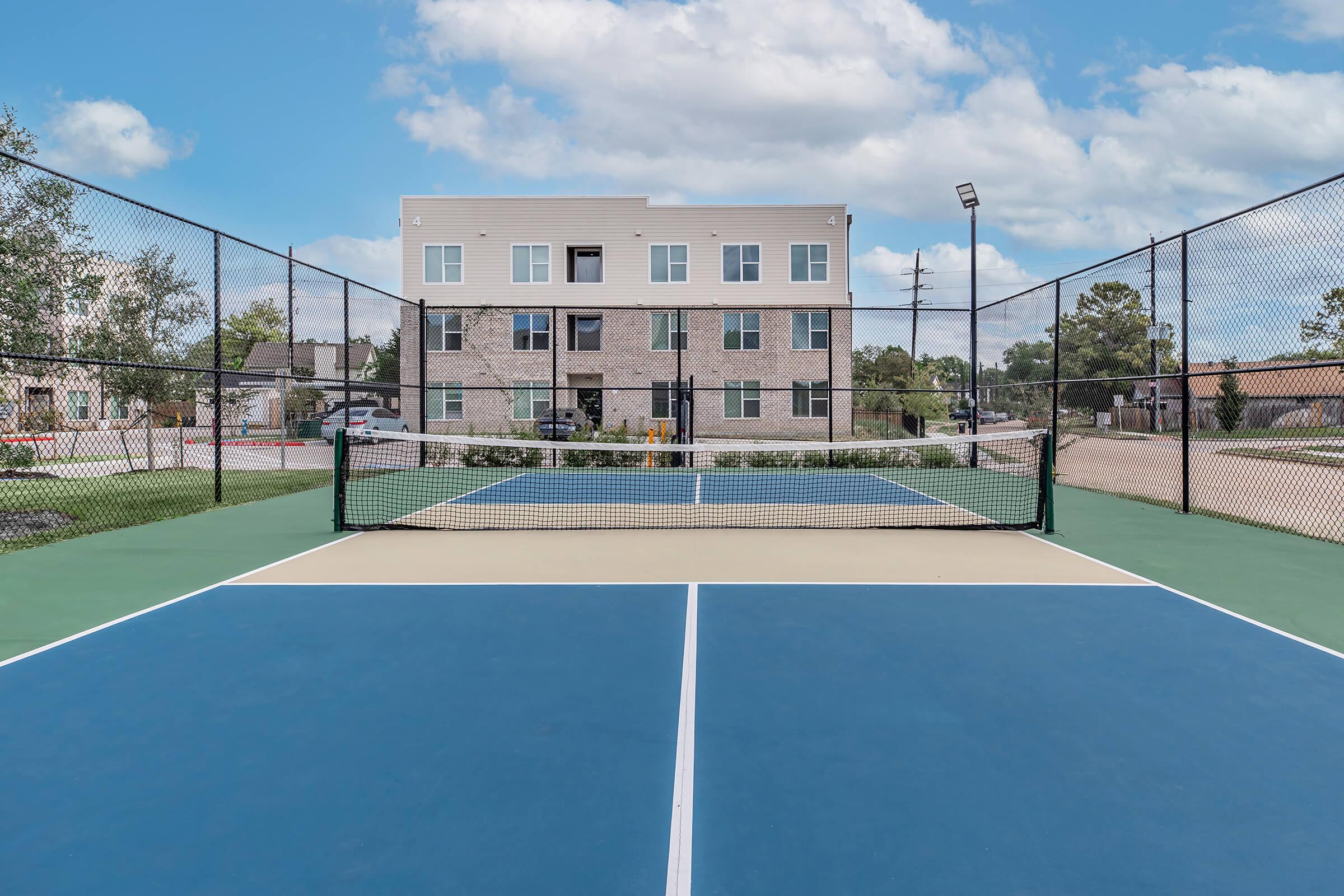 A view of a tennis court with a net in the foreground, featuring vibrant blue and green surfaces. In the background, there is a modern multi-story building surrounded by trees and a parking area. The sky is partly cloudy, adding a bright atmosphere to the scene.