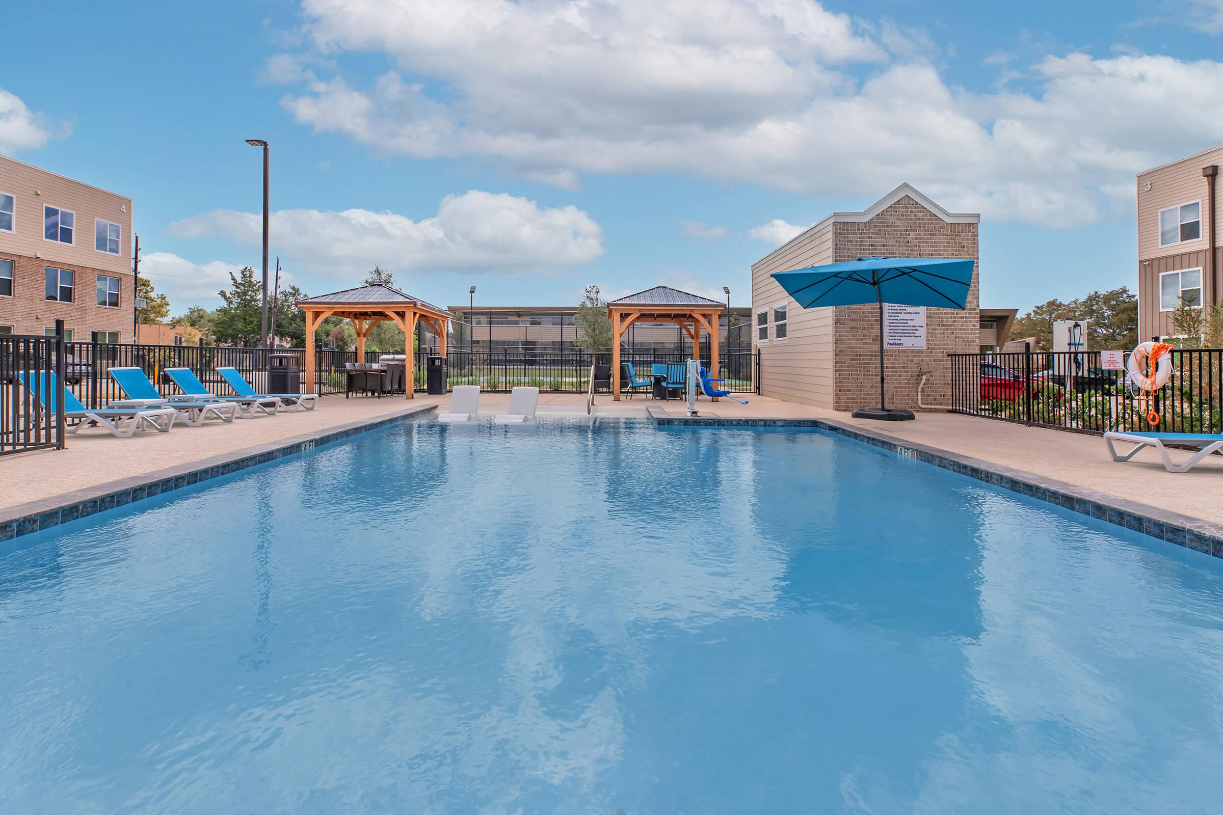 A clear blue swimming pool surrounded by lounge chairs and shaded cabanas. The pool area features a clean, inviting atmosphere with buildings in the background and a bright sky above. Perfect for relaxation and leisure activities.