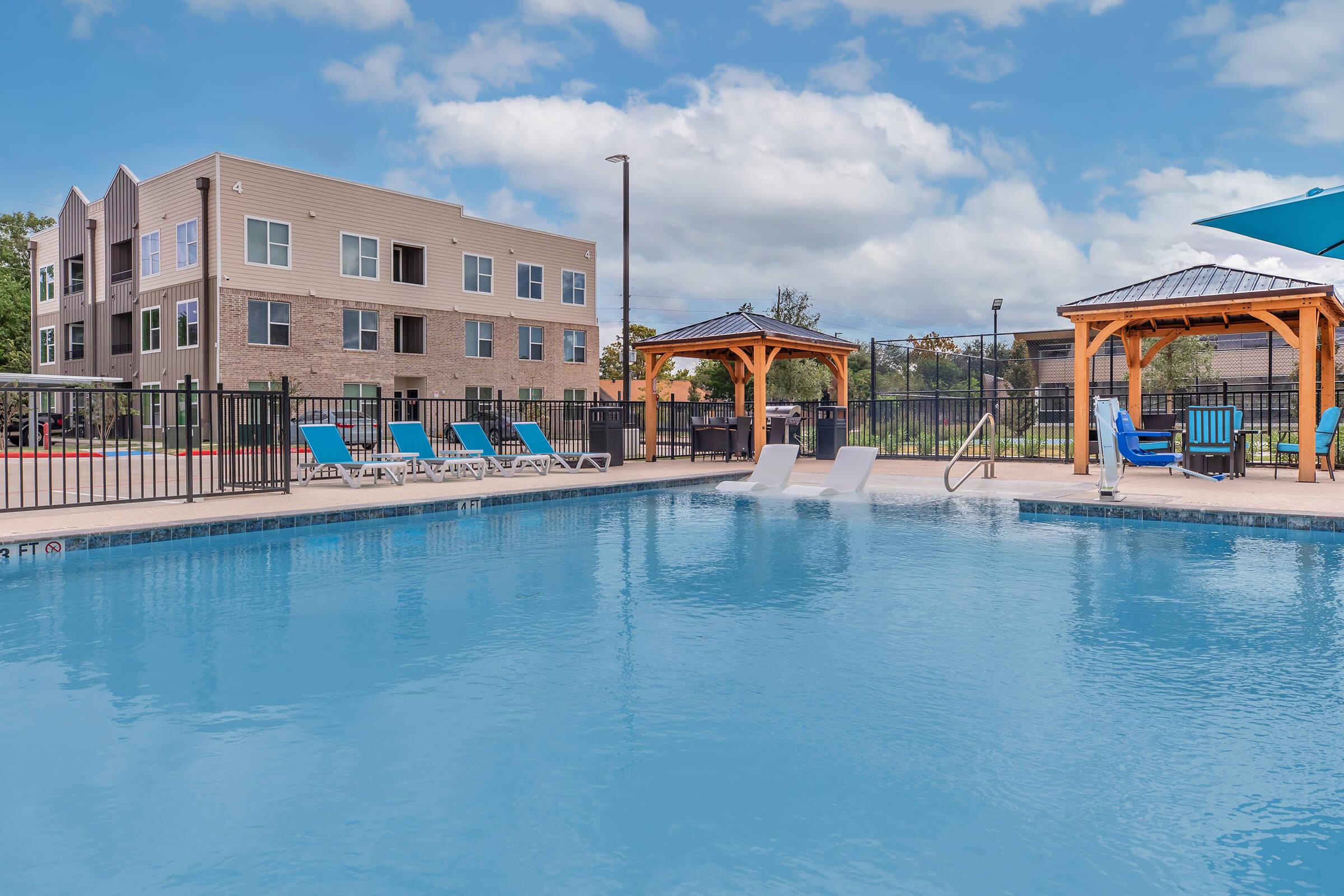 A clear swimming pool with lounge chairs around it, featuring a gazebo and an outdoor seating area. In the background, a modern apartment building is visible under a partly cloudy sky. The scene conveys a relaxed outdoor atmosphere ideal for leisure and recreation.