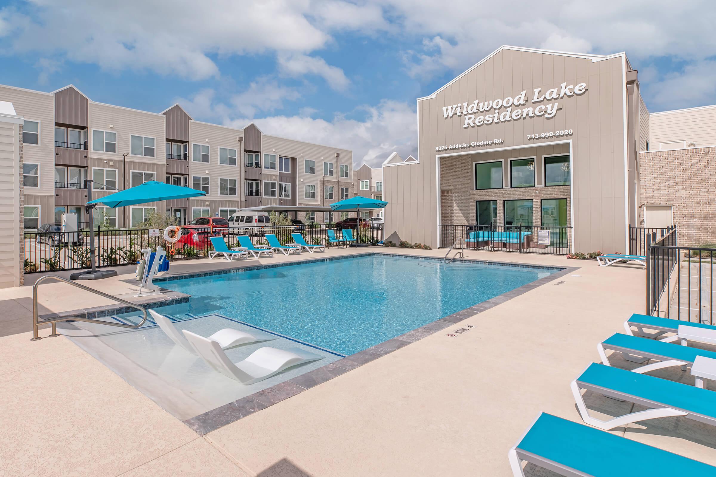 A modern apartment complex featuring a pool area with lounge chairs and umbrellas. The pool is surrounded by a stone patio, and the building has large windows and a welcoming entrance sign that reads "Wildwood Lake Residency." Clear blue skies and a well-maintained landscape enhance the setting.