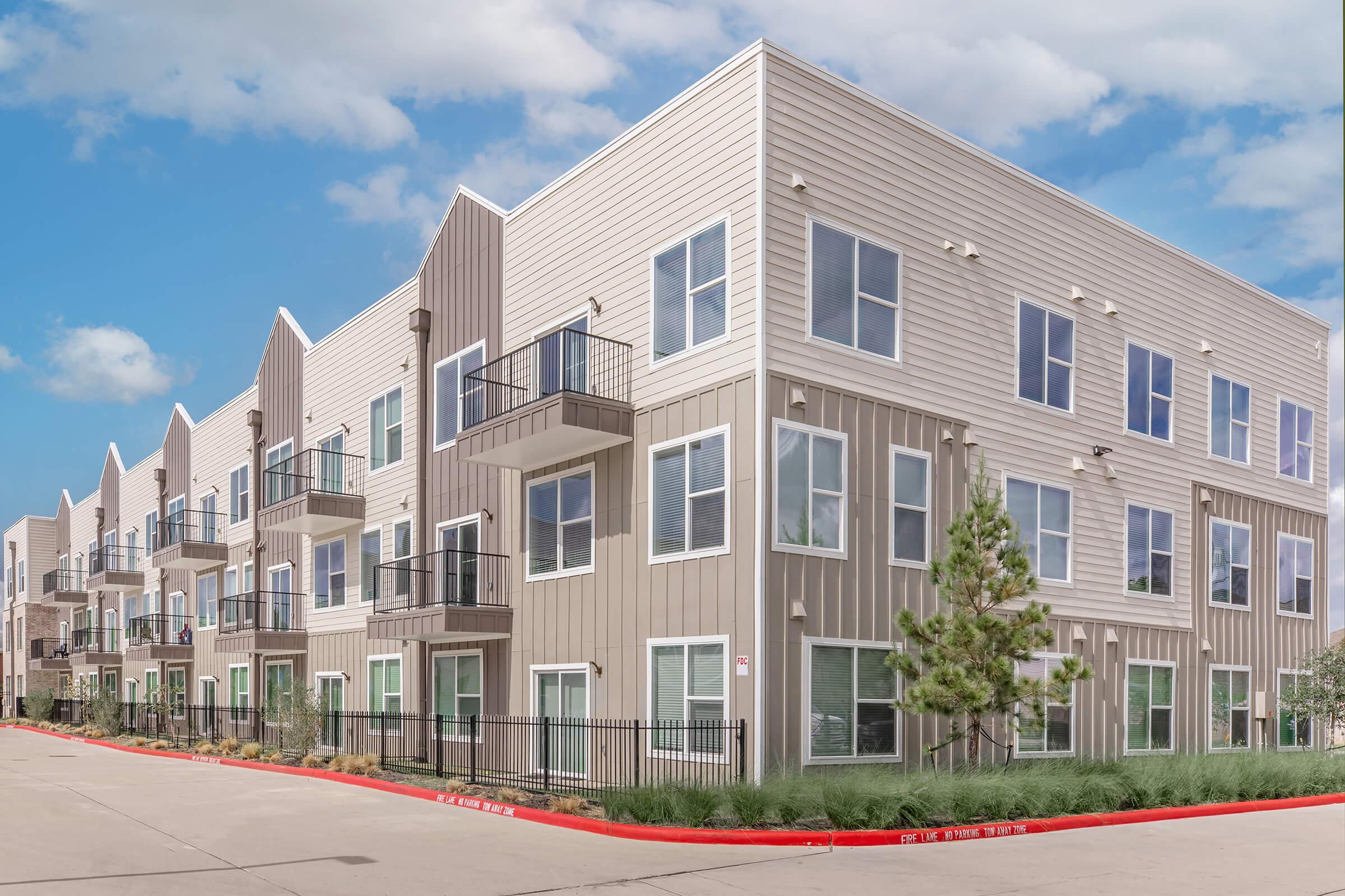 A modern residential building featuring a series of multi-story units with balconies, large windows, and a light-colored exterior. The structure is surrounded by landscaped areas and a paved pathway. The sky above is partly cloudy, enhancing the contemporary architectural design.