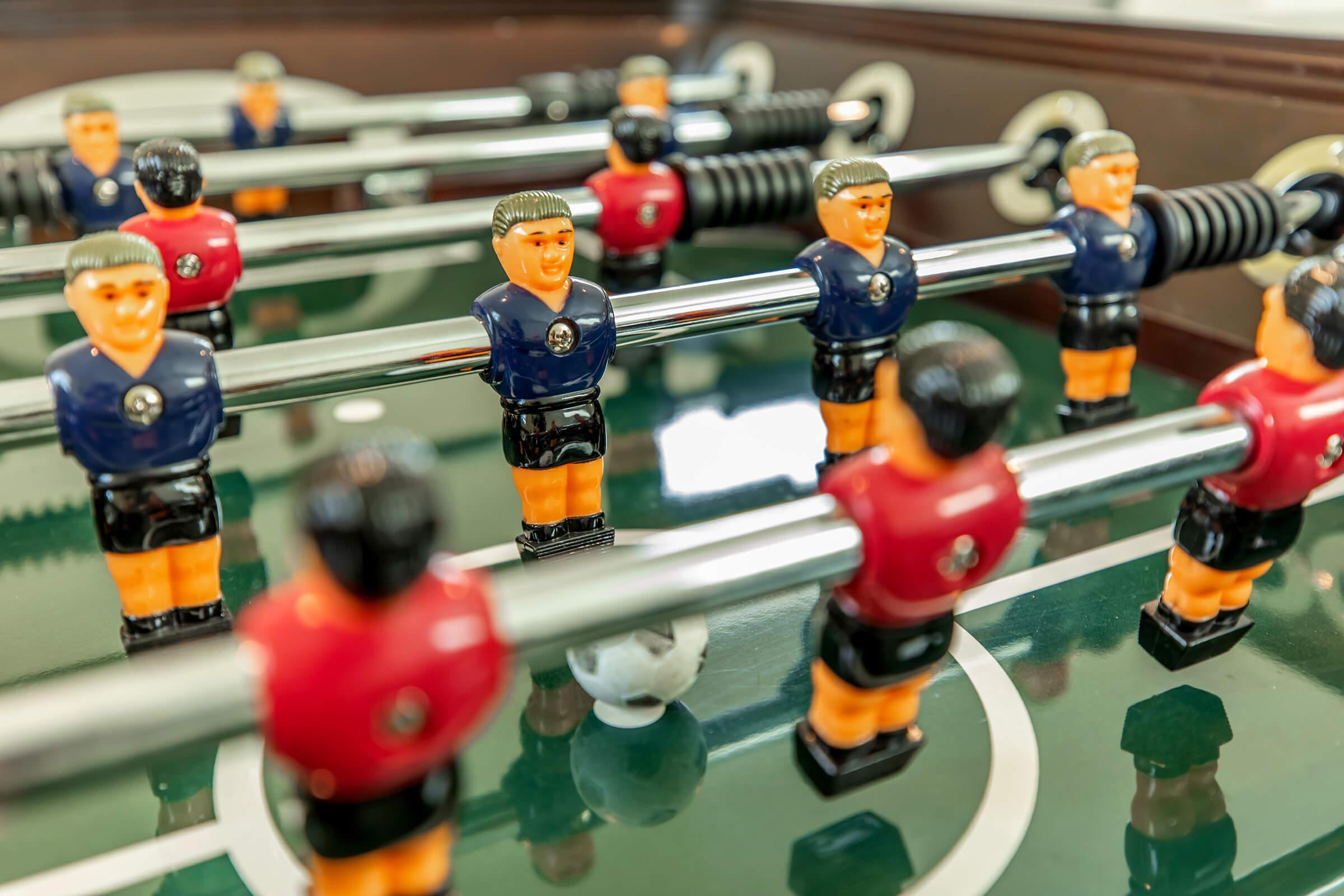 A close-up view of a foosball table with players in blue and red jerseys. The players are positioned around a small white ball on a green playing surface, with metal rods visible. The scene captures the excitement of a foosball game in action.