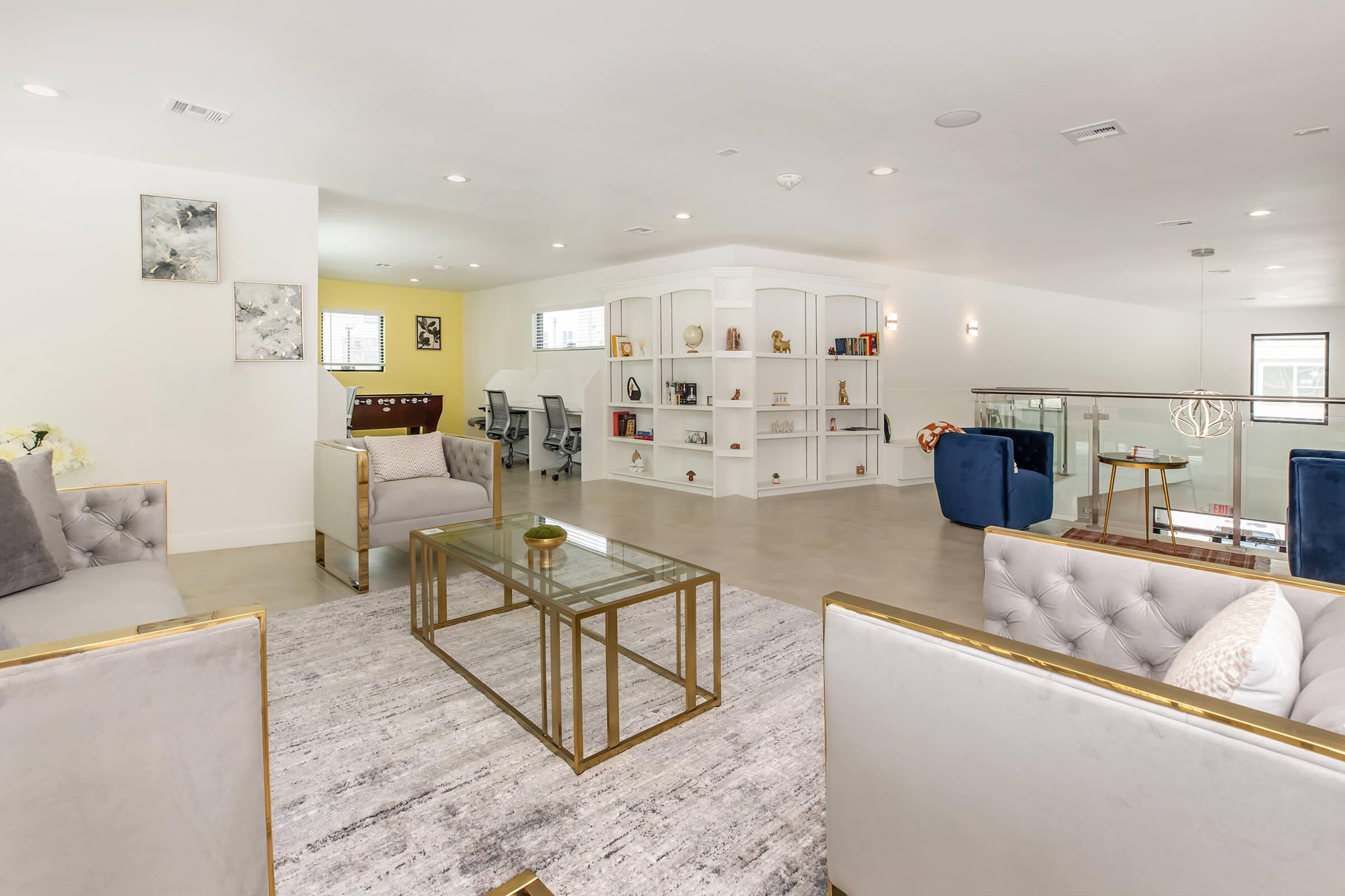 A modern, spacious living area featuring two stylish gray sofas with gold accents, a glass coffee table, and a light-colored area rug. A built-in bookshelf displays decorative items, and a background area includes workstations with chairs. The ambiance is bright and contemporary.