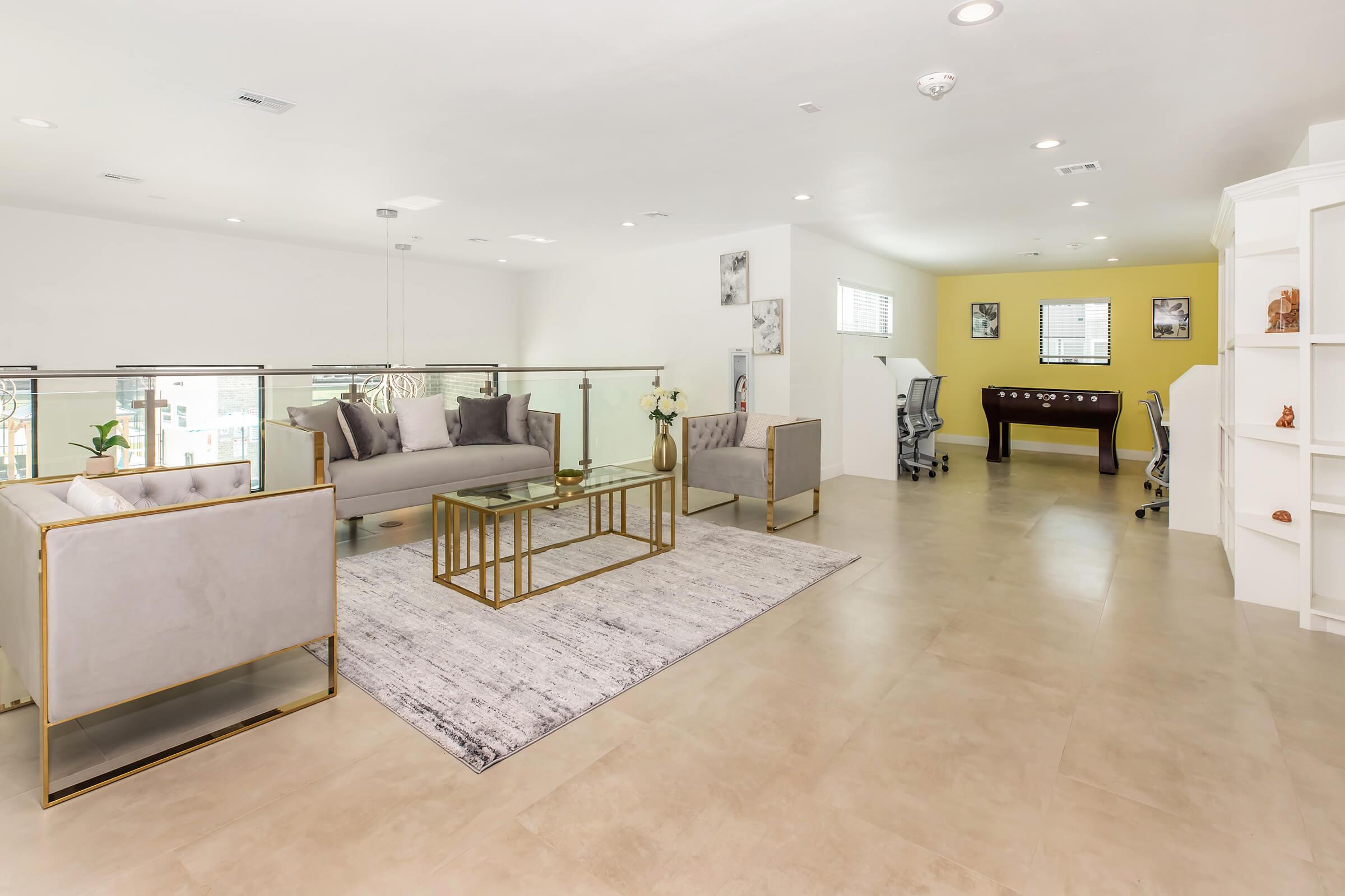 A modern and spacious living area with a light color palette. The room features a gray sofa set with gold accents, a matching coffee table, and a decorative rug. There are elegant chairs and a desk area in the background against a yellow wall. Natural light floods the space through large windows.