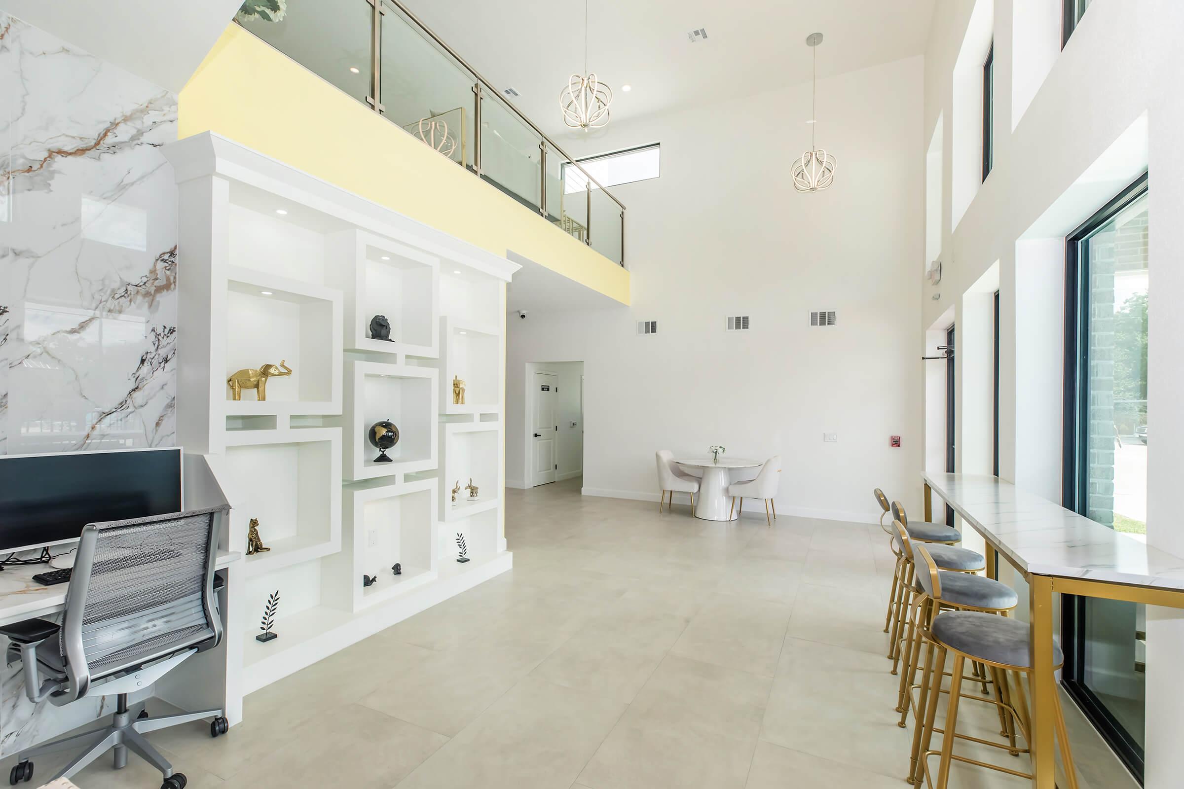A modern, bright lobby featuring a high ceiling with large windows, white walls, and a decorative shelf displaying sculptures. A sleek desk and chair are positioned on one side, while a small dining area with a circular table and chairs is on the other. Decorative lighting adds a stylish touch.