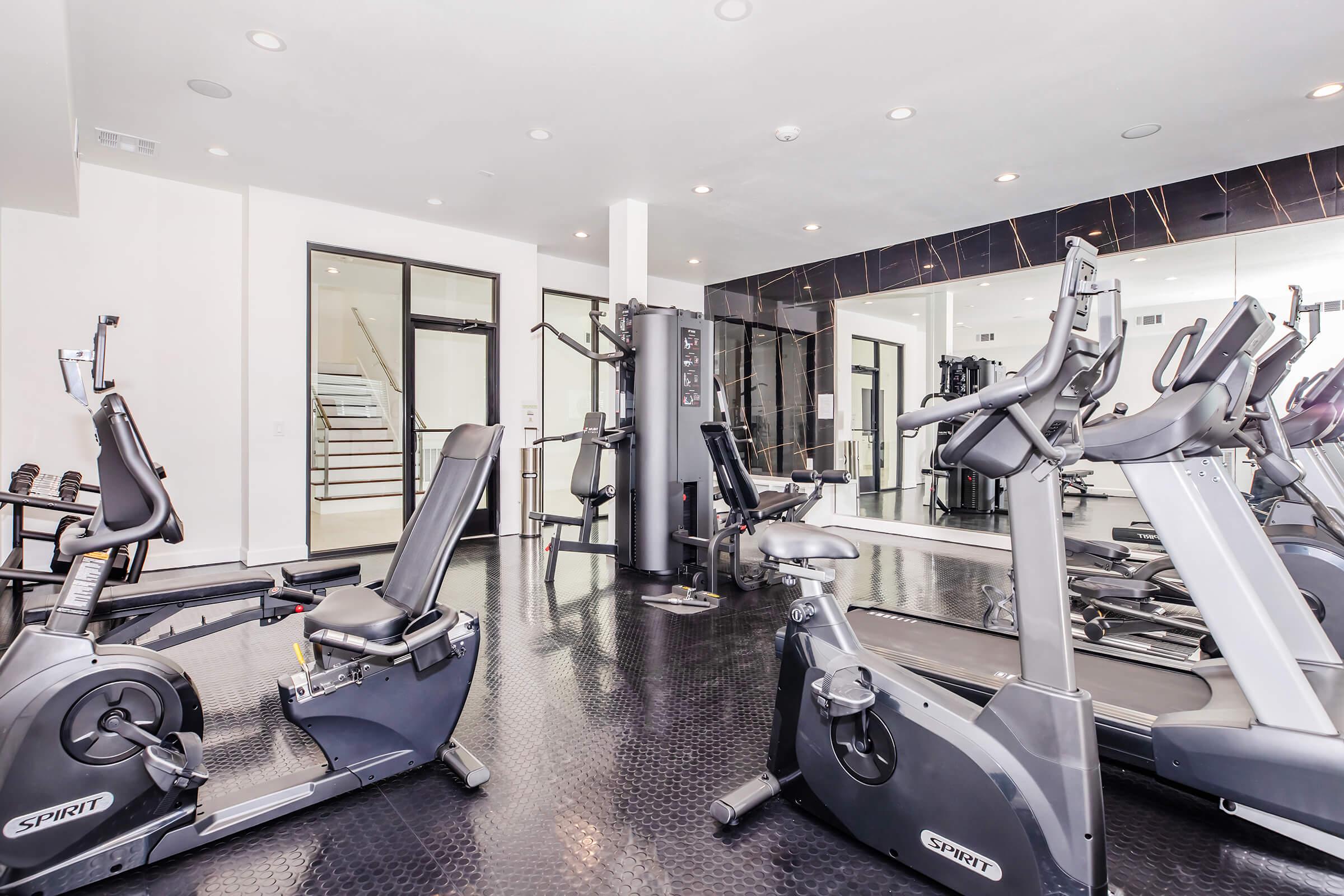 A modern gym interior featuring various exercise equipment, including stationary bikes and elliptical machines. The space is bright with large windows, sleek flooring, and mirrors on the walls, creating a clean and inviting atmosphere for workouts.