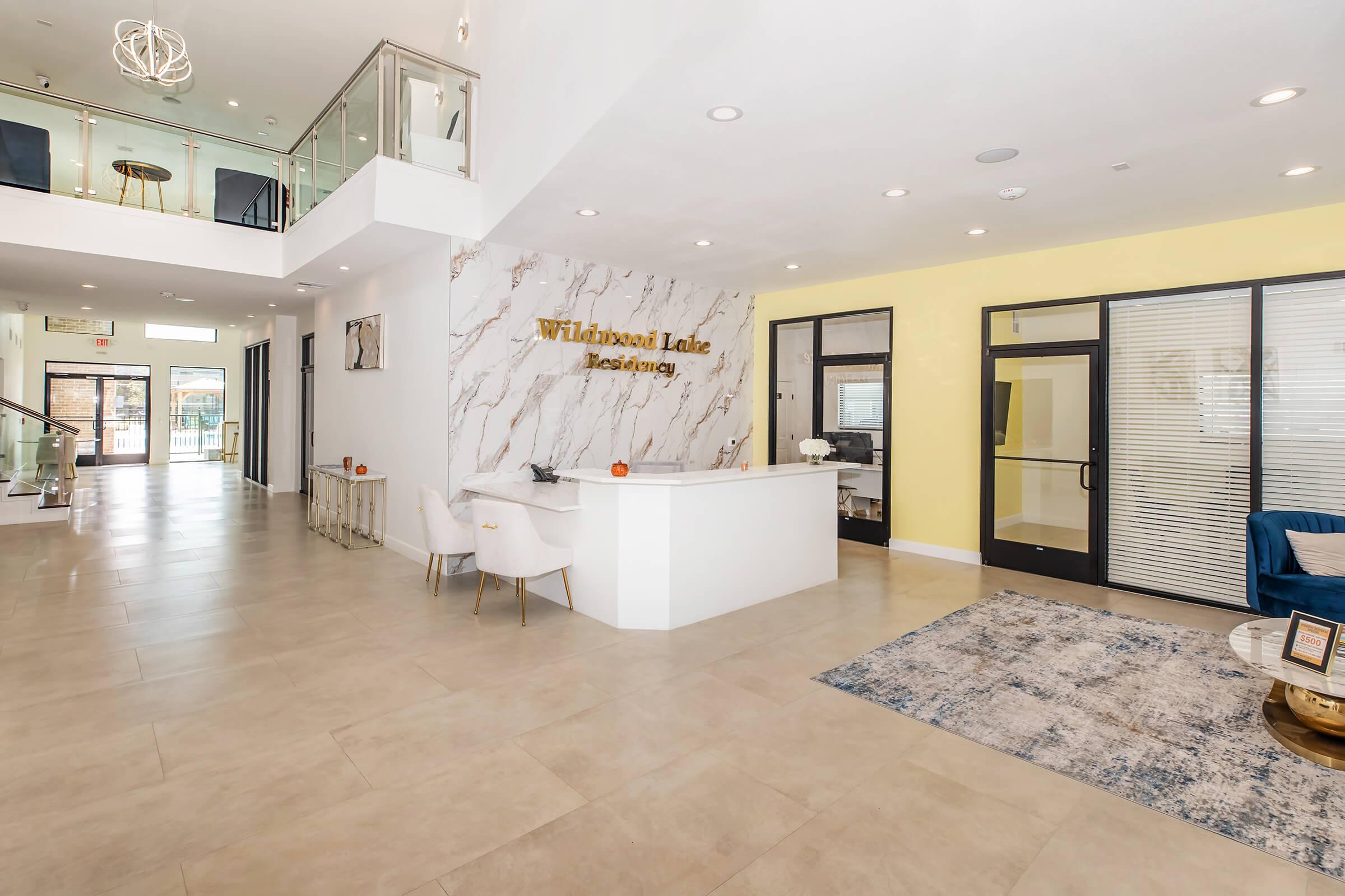 Modern lobby of a leasing office featuring a stylish reception desk with a marble design, surrounded by contemporary seating. The space is brightly lit with large windows and includes decorative elements like a rug and wall signage that reads "Wildwood Lake Leasing."
