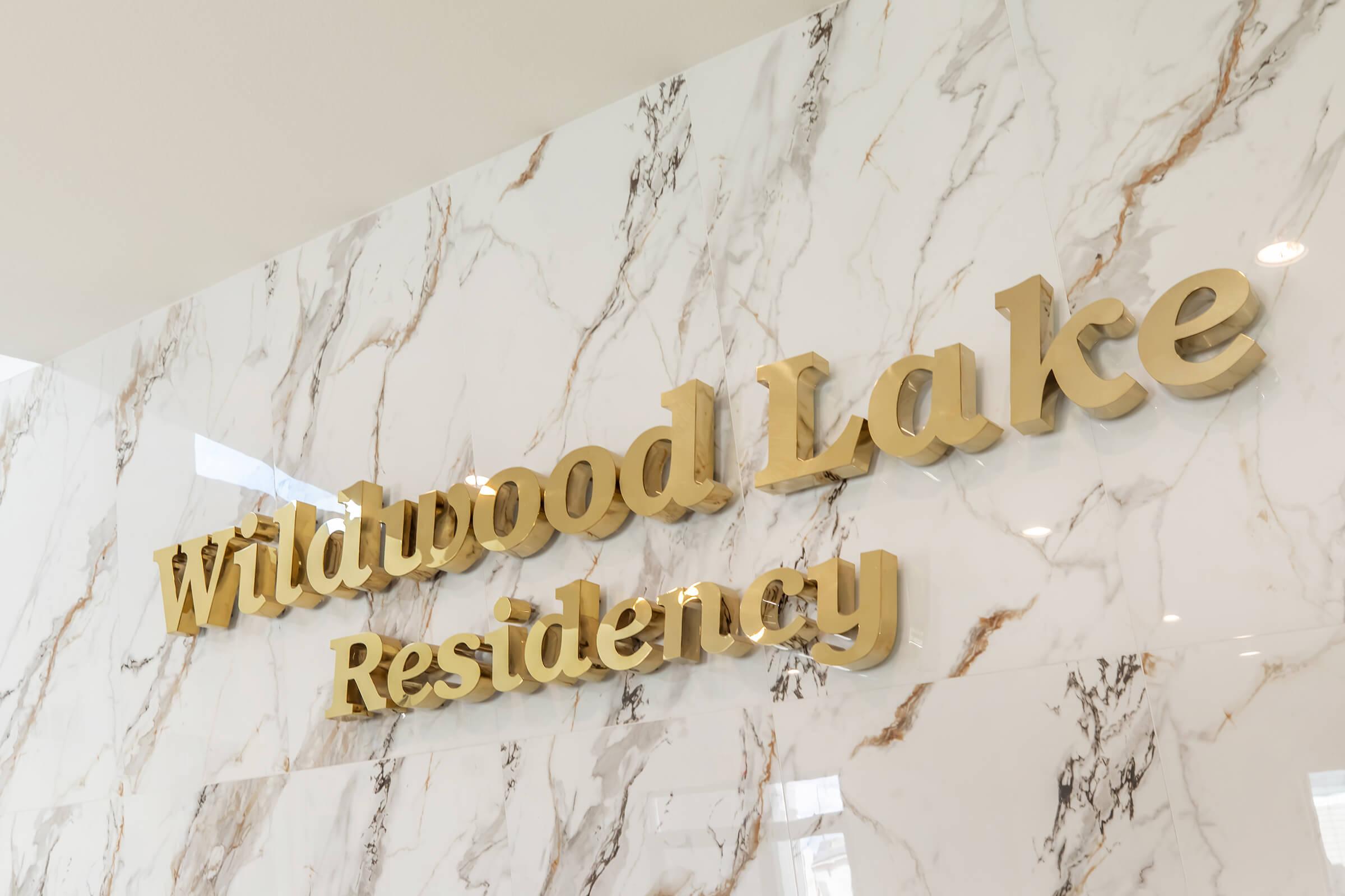 Gold lettering of "Wildwood Lake Residency" mounted on a marble wall, featuring a smooth and elegant design. The marble has a white background with subtle marbling patterns, enhancing the sophisticated aesthetic of the space.