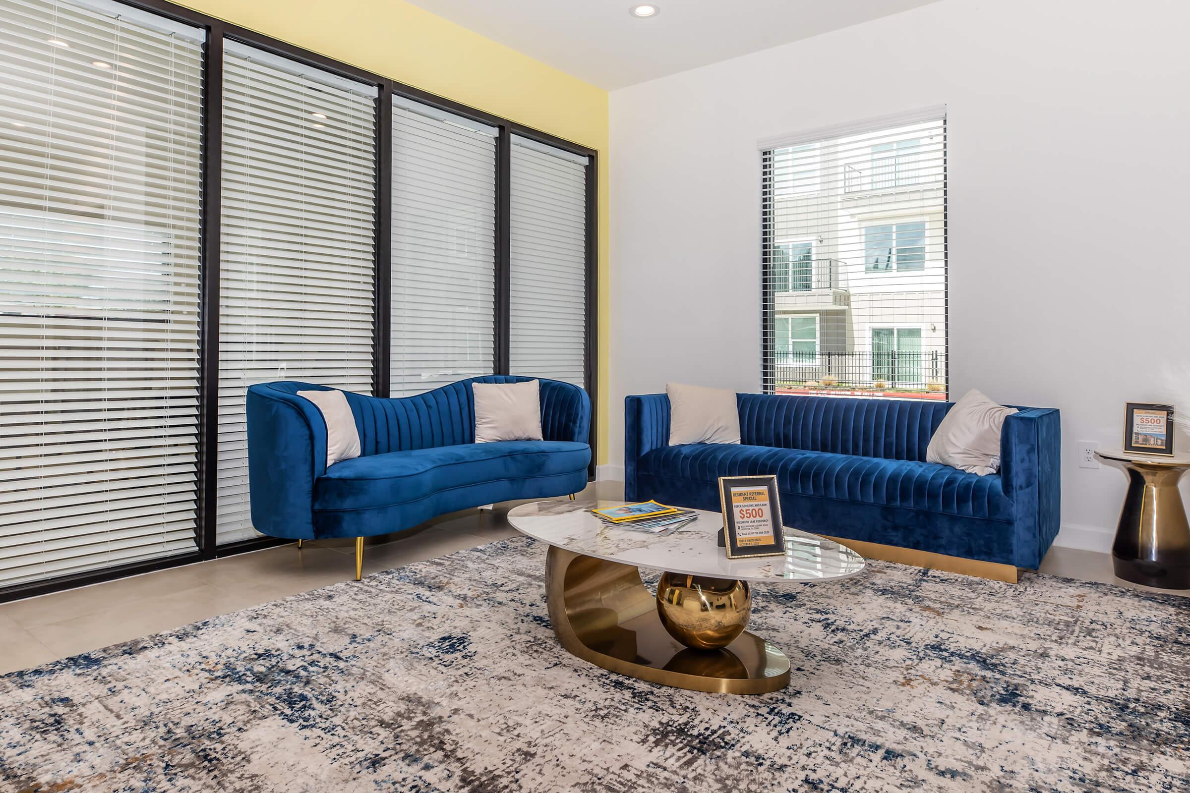 A modern living room featuring two navy blue velvet sofas with light-colored cushions, a stylish round coffee table with a gold base, and a textured area rug. Large windows with horizontal blinds allow natural light into the space, and a vibrant yellow wall adds a pop of color.
