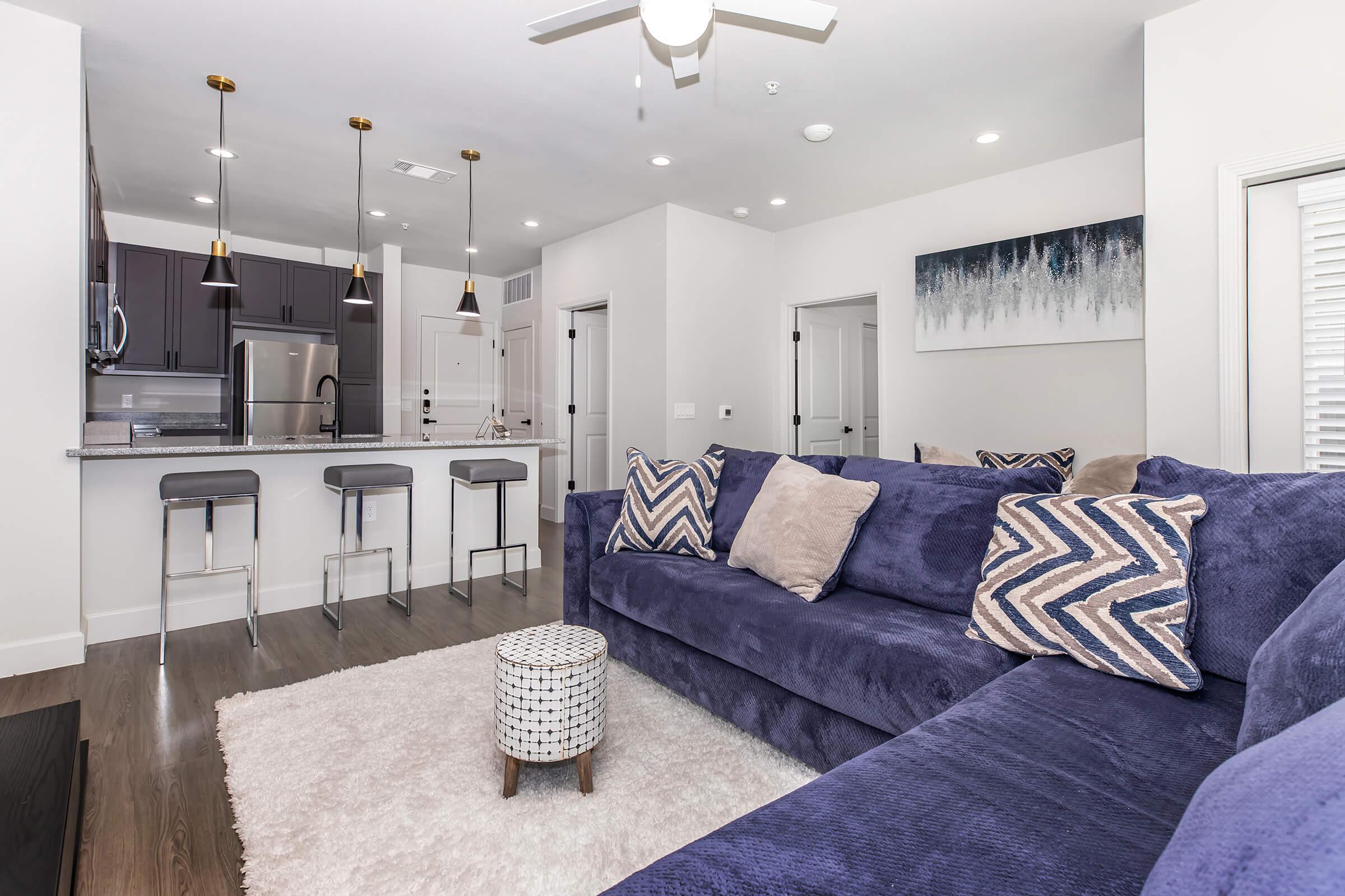 A modern living room featuring a plush navy blue sofa with decorative pillows, a round coffee table, and a soft area rug. The open layout includes a kitchen with bar seating, stainless steel appliances, and stylish pendant lights. Light-colored walls and a framed landscape artwork enhance the contemporary design.