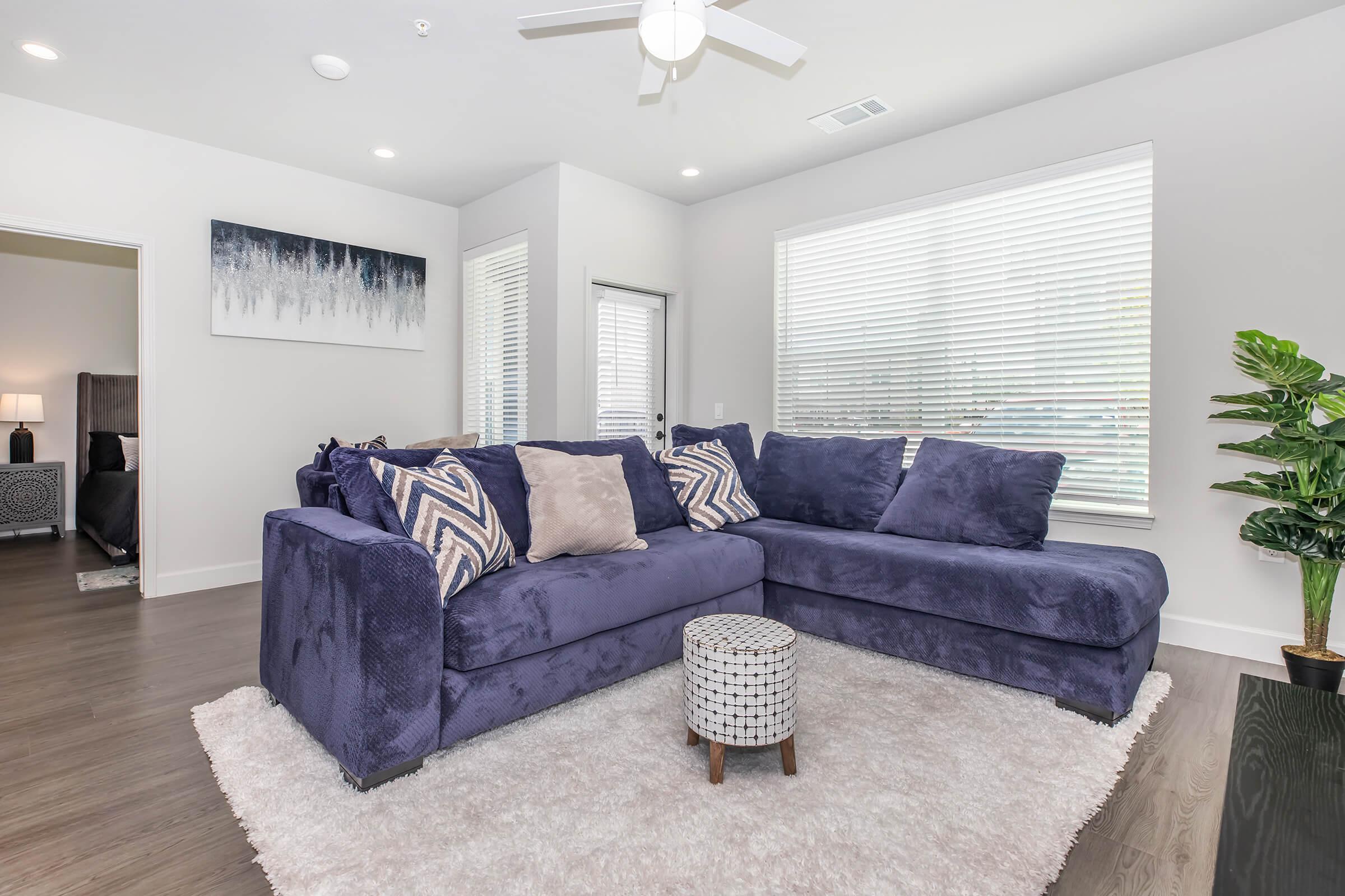 A cozy living room featuring a large purple sectional sofa with decorative pillows, a small round coffee table, and a plush area rug. Sunlight filters through the blinds on the windows, enhancing the modern decor, which includes a leafy plant and a wall art piece in the background.