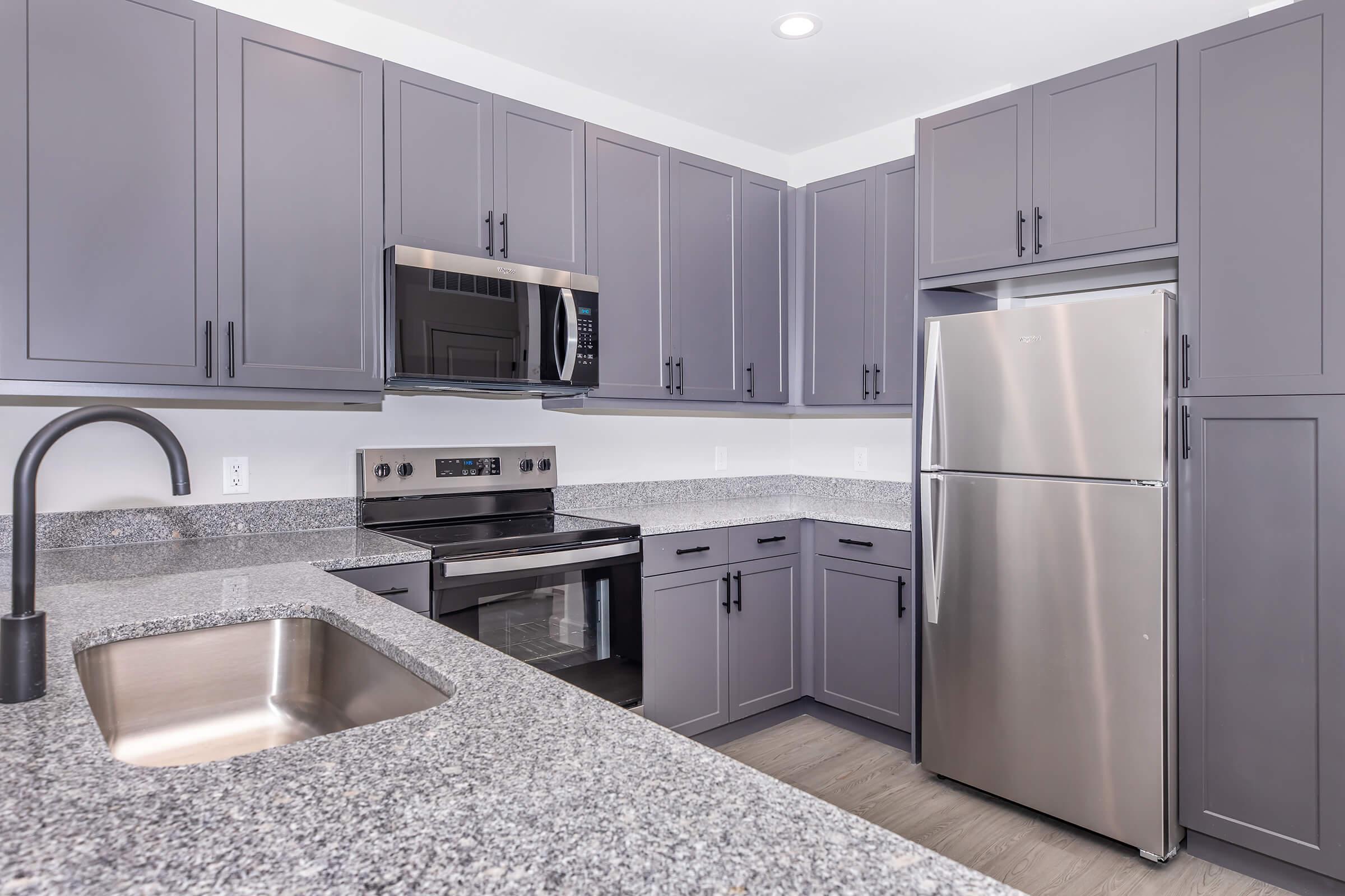 A modern kitchen featuring gray cabinets, stainless steel appliances including a microwave, oven, and refrigerator, along with a stainless steel sink and a granite countertop. The design is contemporary, with ample storage space and bright lighting, creating a welcoming atmosphere.