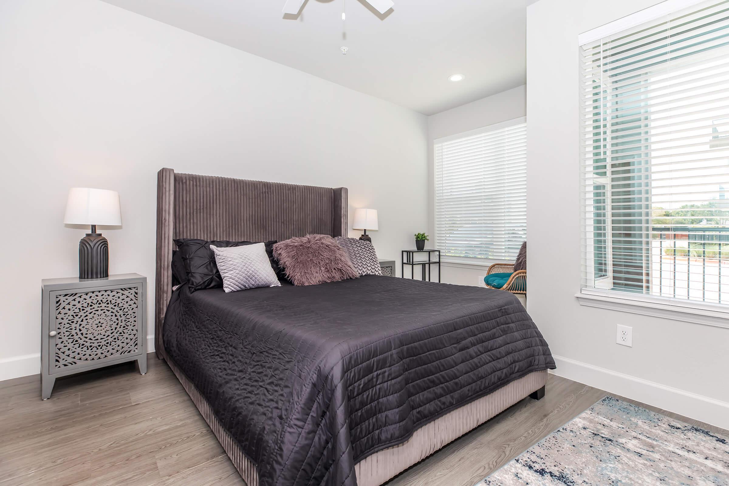 Modern bedroom featuring a queen-sized bed with a dark coverlet and decorative pillows, accompanied by bedside tables with lamps. A window with blinds allows natural light into the room, and a chair is visible in the corner. The floor is wooden, and a colorful rug adds warmth to the space.