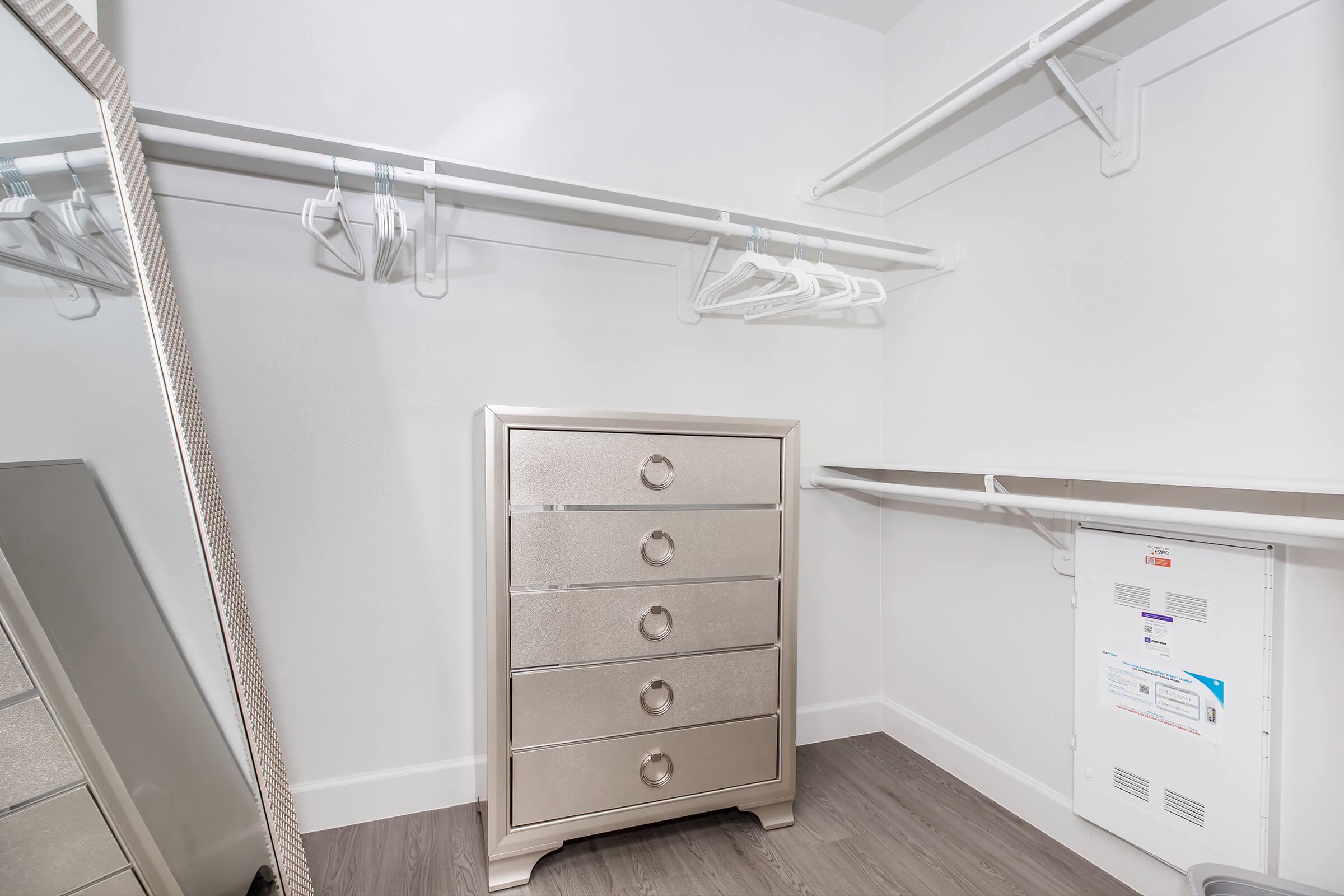 A well-organized closet featuring a stylish silver dresser with five drawers, hanging rods with empty hangers, and a large mirror reflecting the space. The walls are painted white, and the flooring is a light wood finish, emphasizing a clean and modern aesthetic.