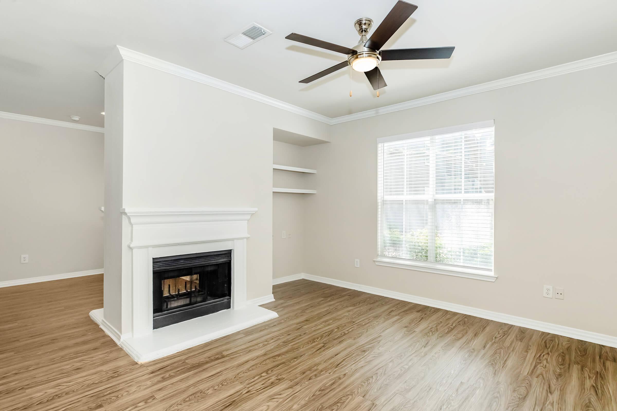 Spacious living room featuring a white fireplace, ceiling fan, and large window with blinds. Light-colored walls and hardwood flooring create a bright, airy atmosphere. Built-in shelves are visible on one side of the room.