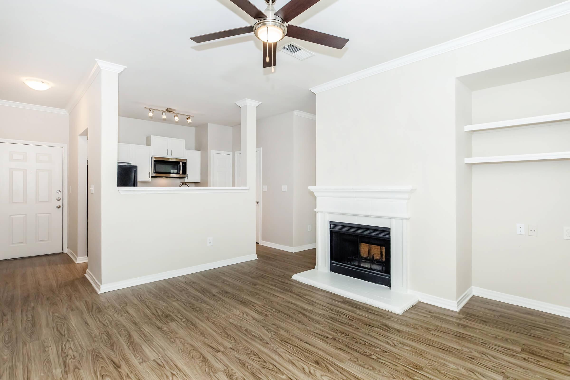 A bright, spacious living room featuring a ceiling fan, a cozy fireplace, and light-colored walls. The room has hardwood-like flooring and is open to a small kitchen area in the background. There are built-in shelves on one side and the entrance doorway is visible.