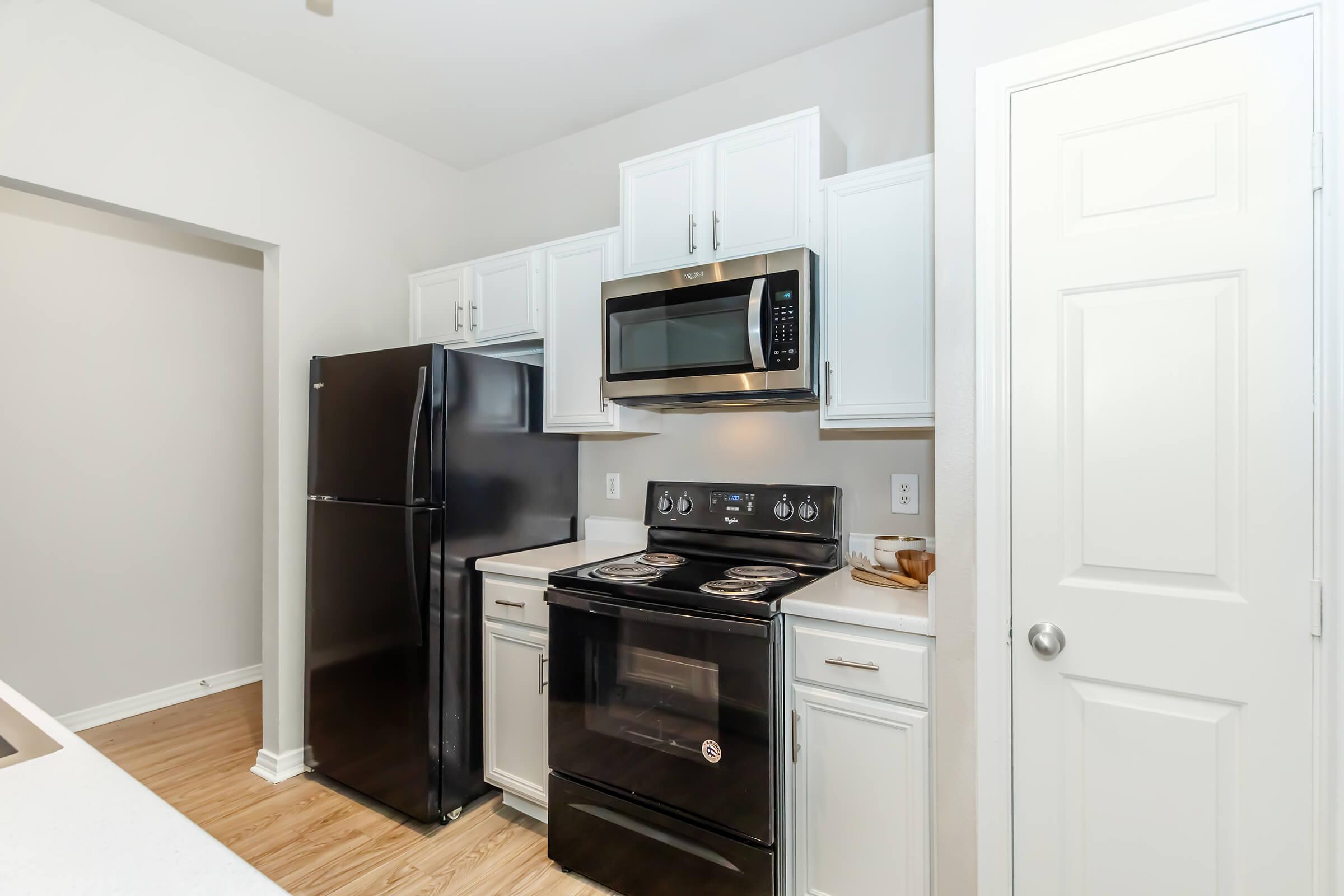 Modern kitchen featuring white cabinets, a black refrigerator, and an electric stove. A microwave is mounted above the stove. Light wood flooring complements the space, and thereās a door on the right leading to another area. The overall design is clean and contemporary.