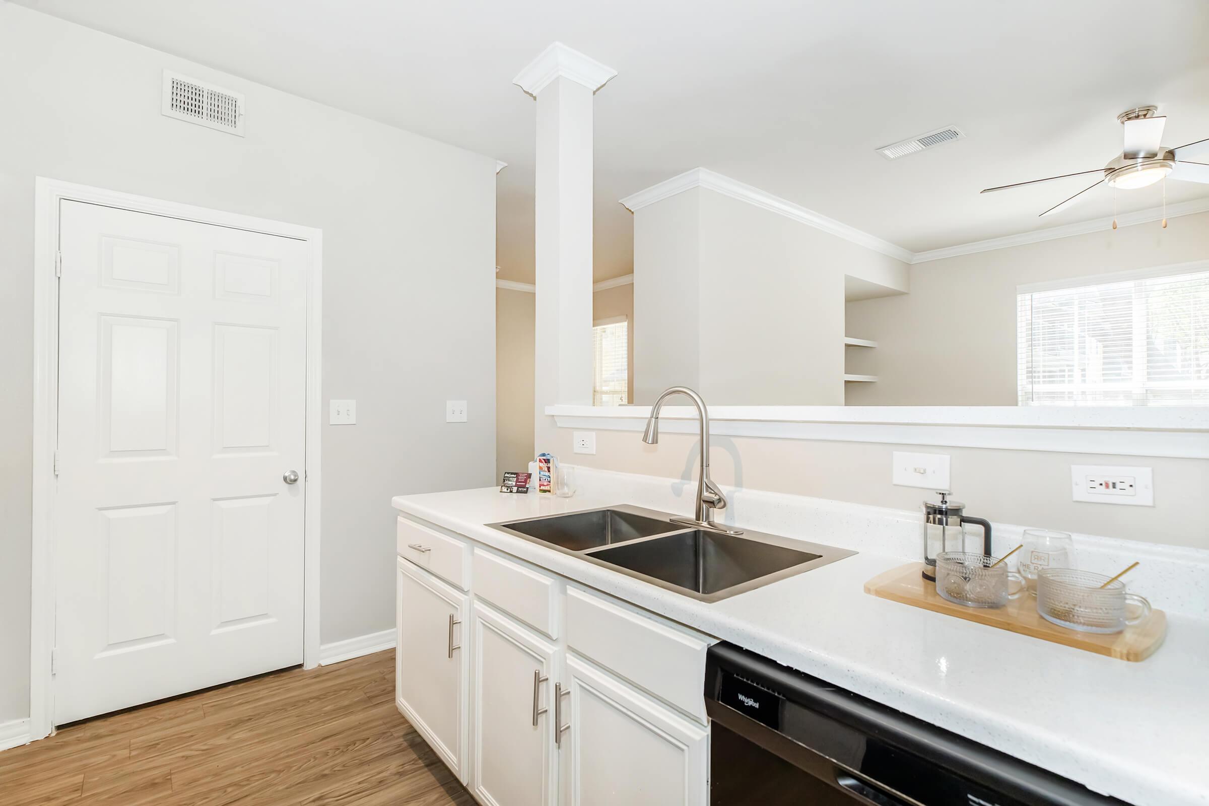 A modern kitchen featuring a dual sink, white cabinetry, and a dishwasher. Natural light flows in from a nearby window. The kitchen is painted in neutral tones, with a small shelf visible in the background and a door leading to another room. The flooring is a warm wood finish.