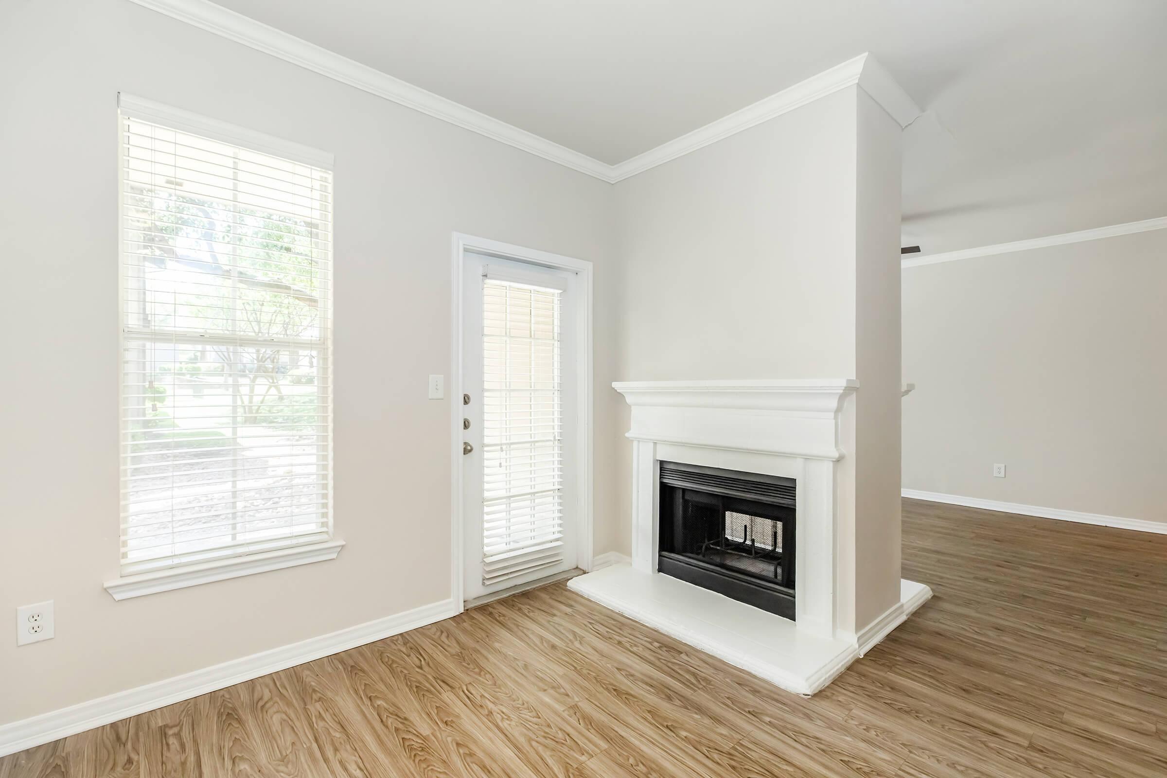 A bright, empty living room featuring a corner fireplace with a white mantel, light-colored walls, and large windows with blinds. There is a door leading outside, and the floor is made of light hardwood. The overall space feels open and airy, with ample natural light.