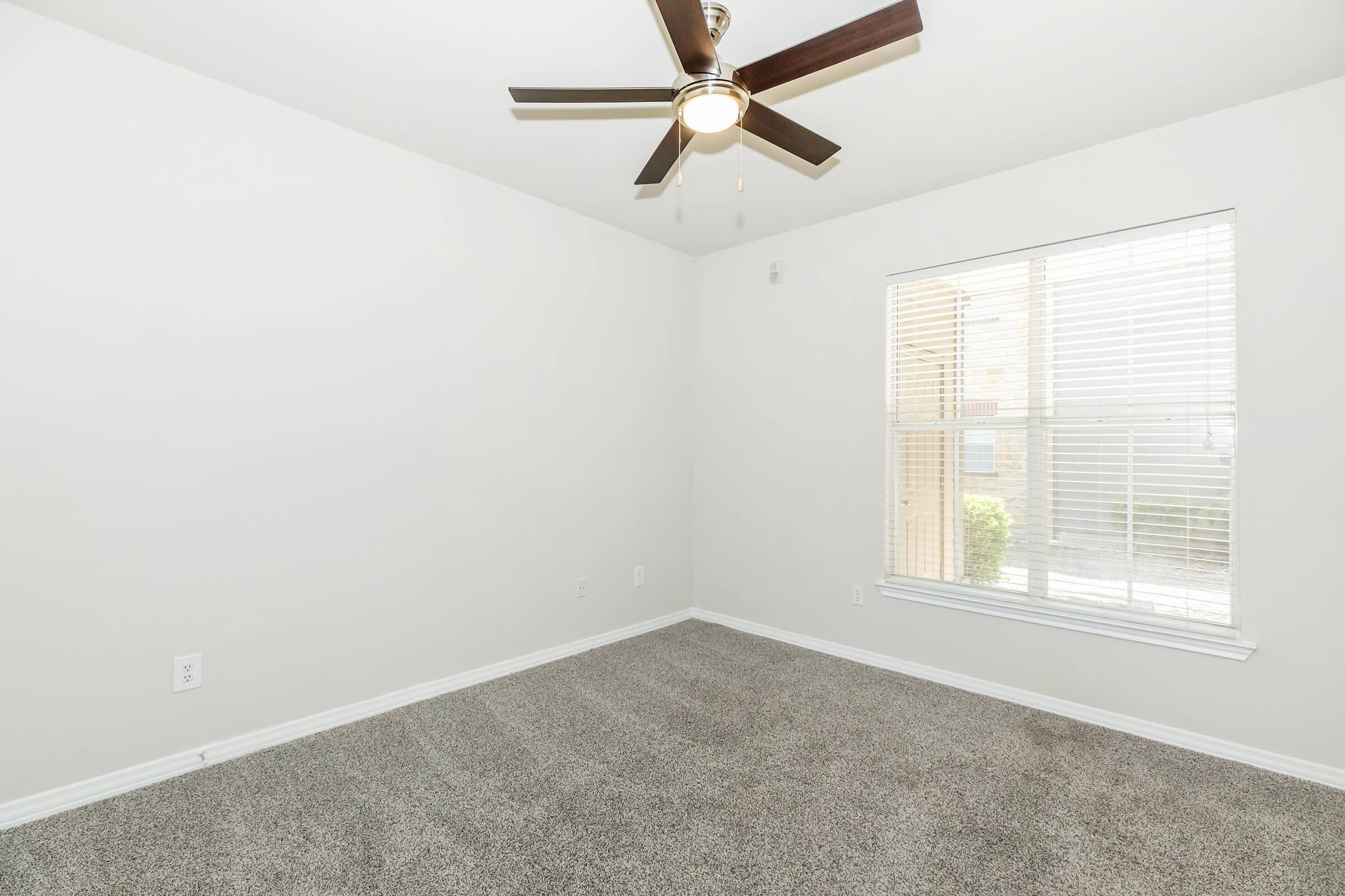 Empty room with light beige walls and a light brown carpet. A ceiling fan with wooden blades is visible. There is a window with white blinds, allowing natural light to enter, and a view of greenery outside. The room appears tidy and spacious, lacking any furniture or decorations.