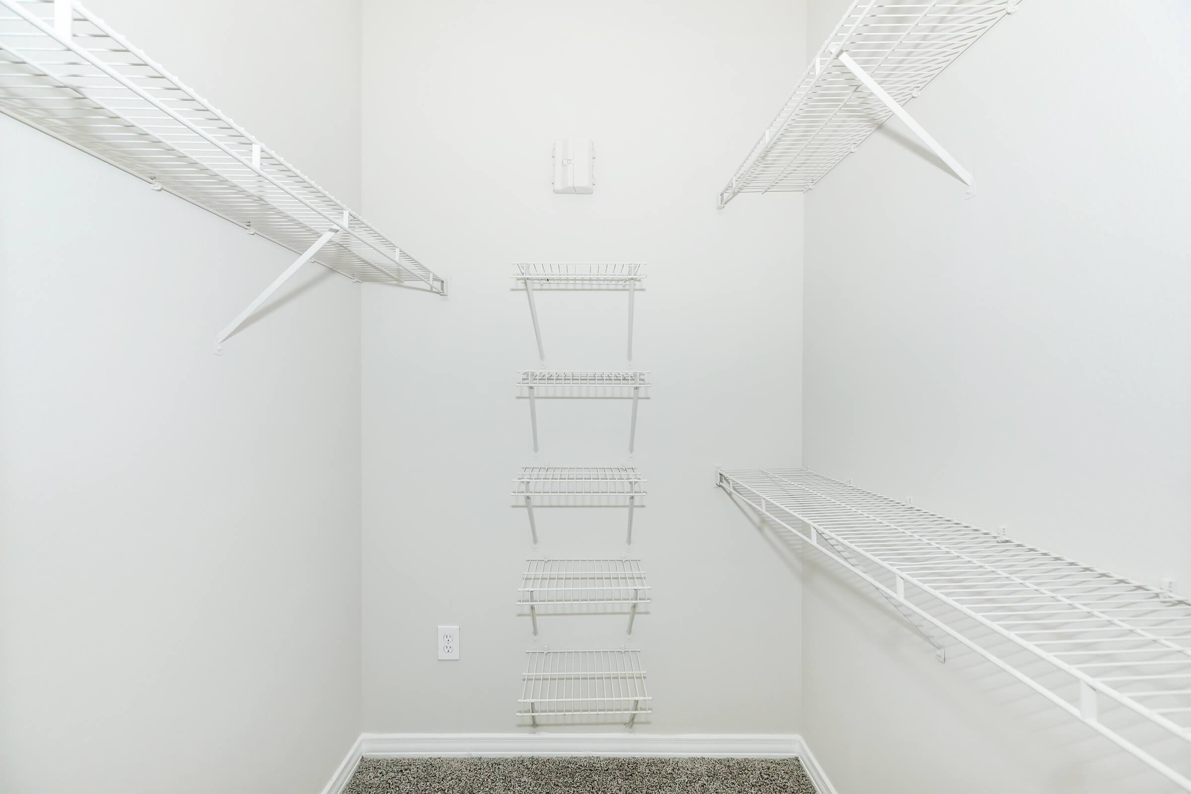 An empty closet with white wire shelving mounted on the walls. The shelves are arranged at various heights, creating an organized space for storage. The walls are painted in a light color, and the floor is covered with a soft gray carpet.