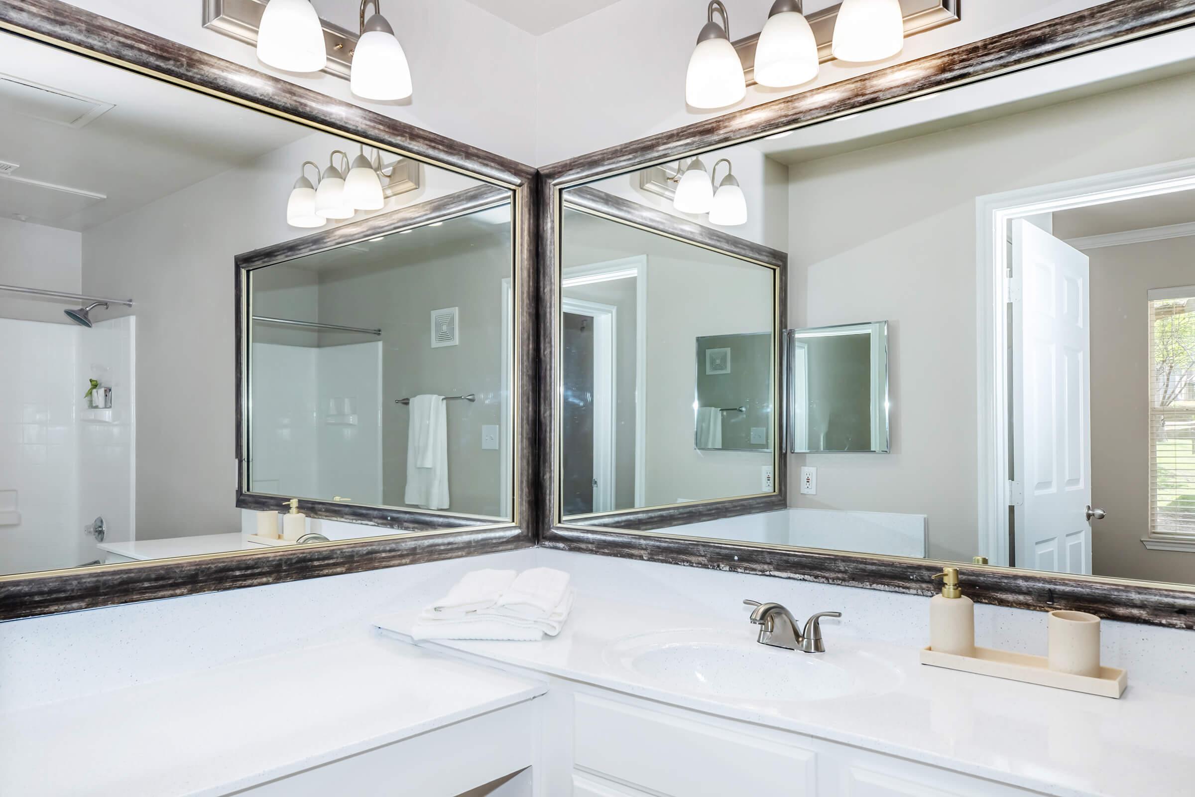 A modern bathroom with a white countertop, dual mirrors framed in dark metal, and decorative items. The space features bright lighting from above and a shower area visible through a frosted glass door. A neatly folded towel and a small decorative tray are placed on the countertop.